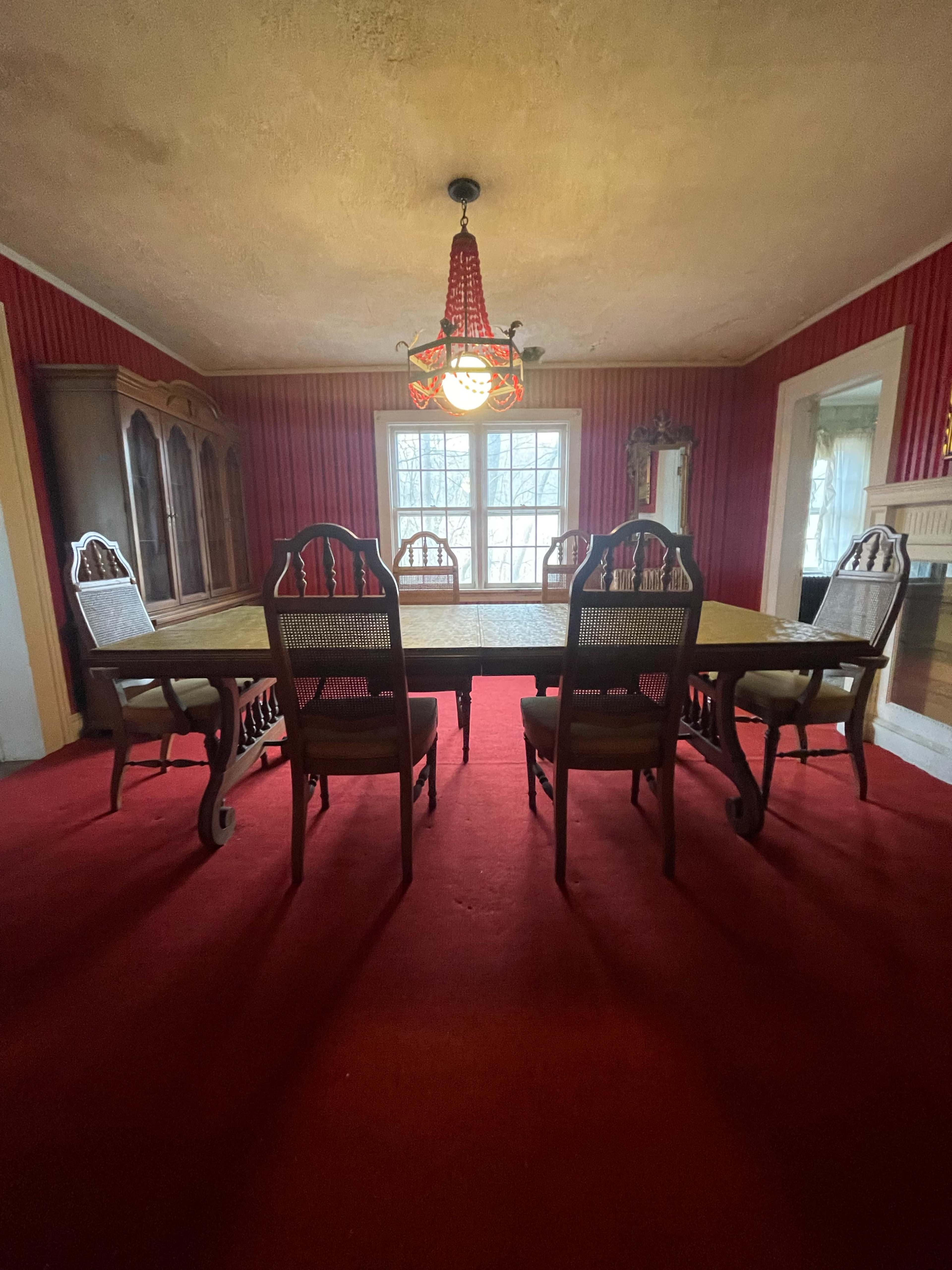 A dining room with a large table surrounded by wooden chairs, red carpet flooring, and a window framed by white curtains.