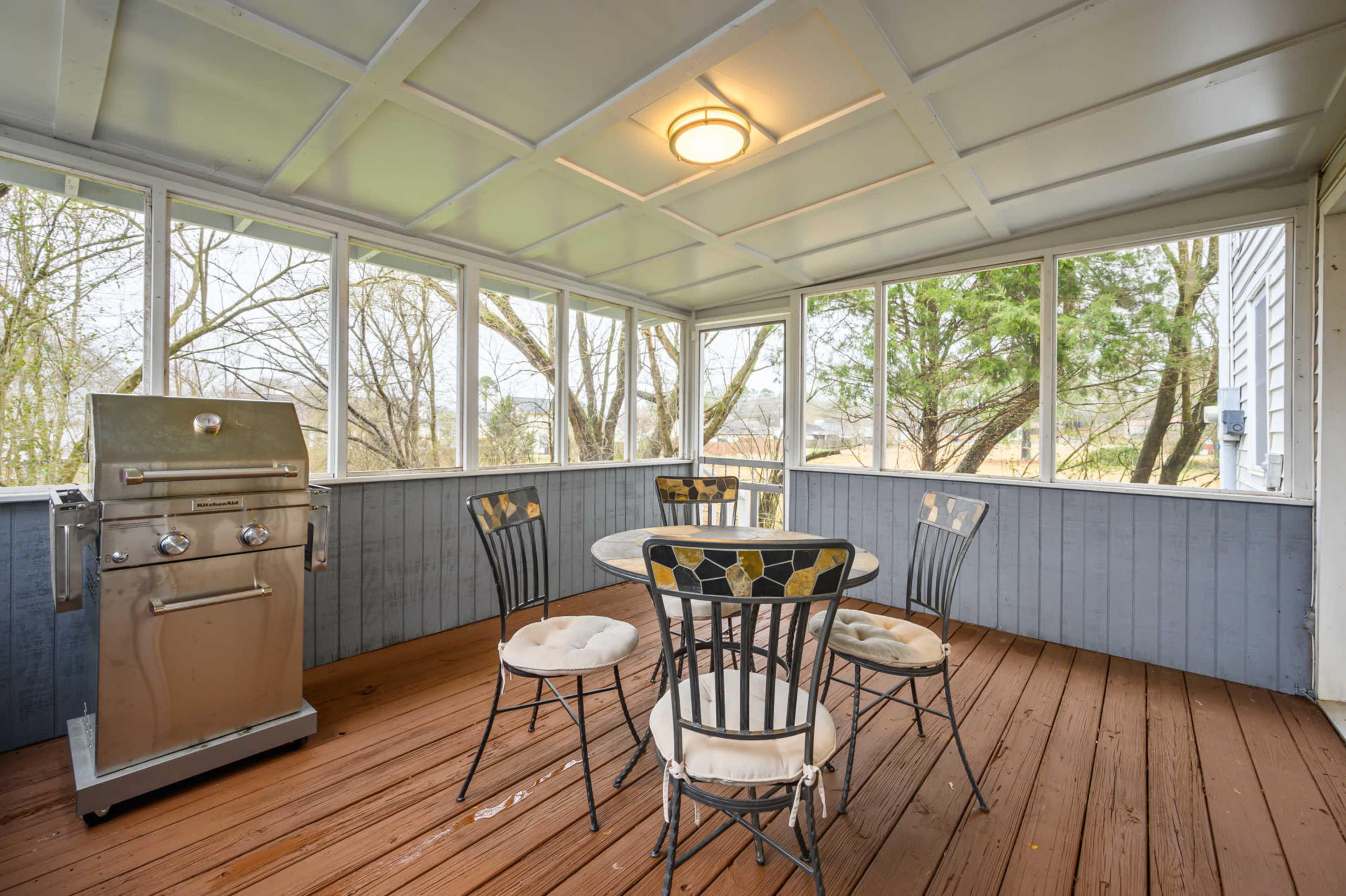 A screened porch features a table with four chairs and a grill in one corner, surrounded by trees visible through the large windows.