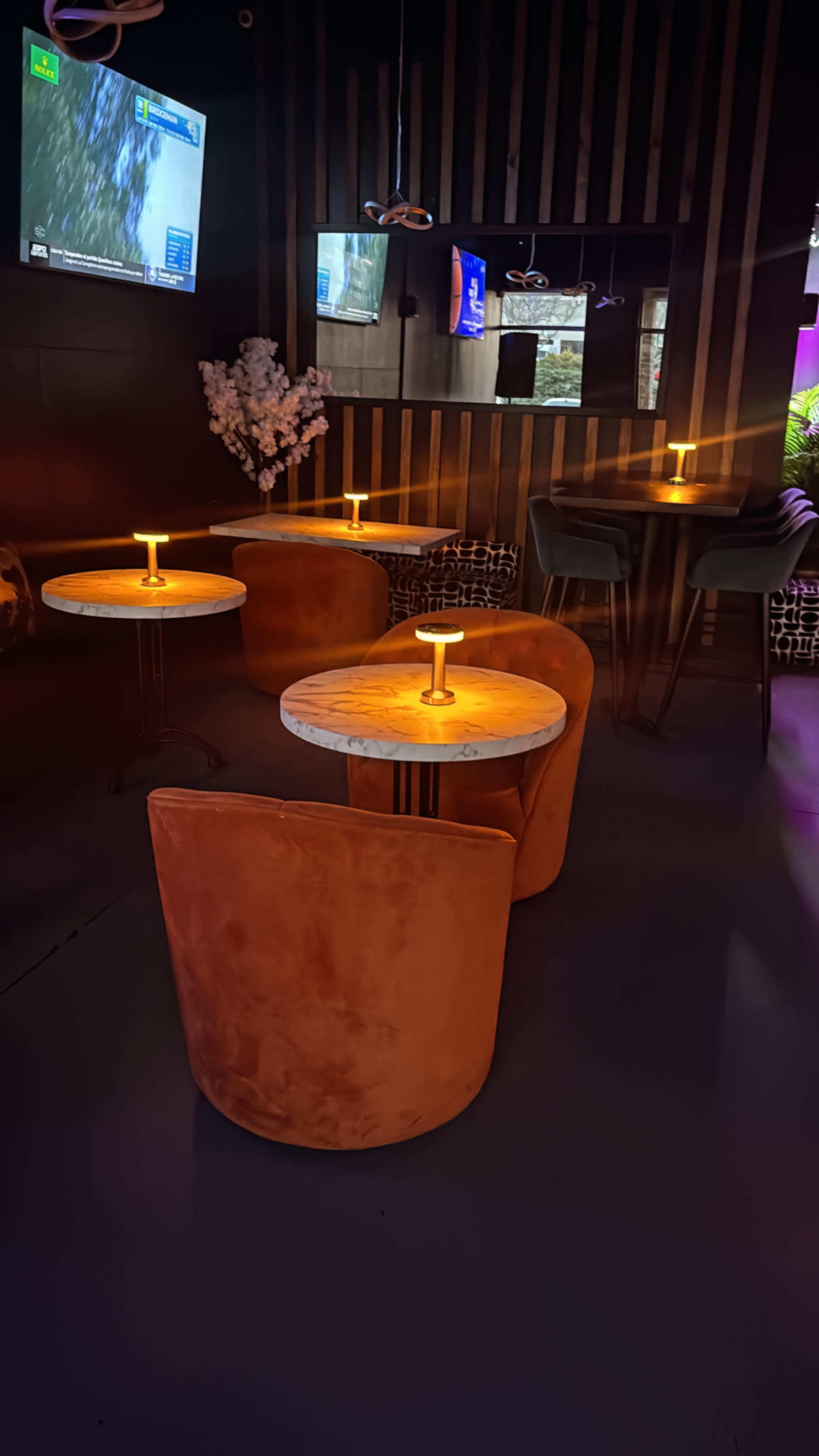 The image shows a dimly lit lounge area with four circular tables, each accompanied by a single illuminated candle, and orange velvet chairs positioned around them.