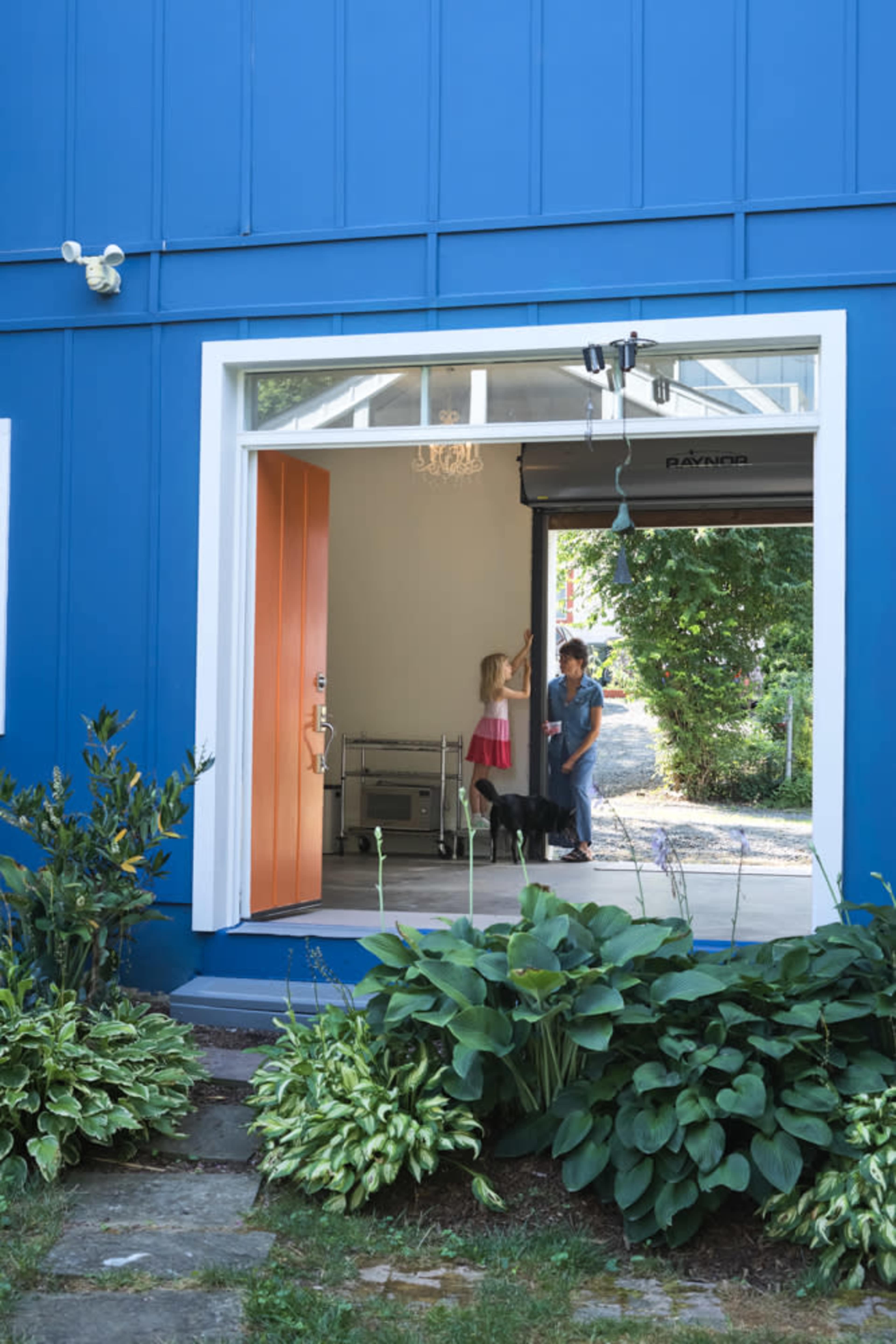 A woman and a girl interact inside a brightly colored blue house with an open doorway, while a dog stands nearby.