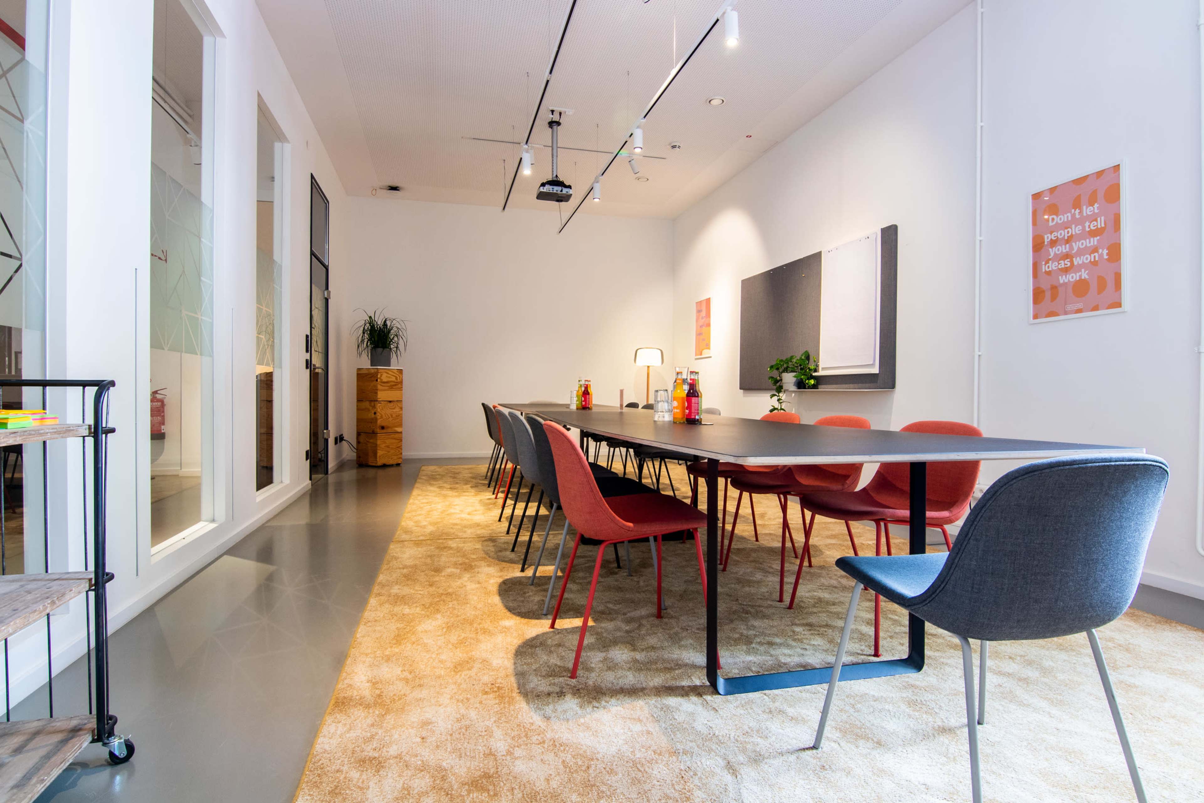 A modern conference room features a long table surrounded by red and gray chairs, with a projector on the ceiling and decorative elements on the walls.
