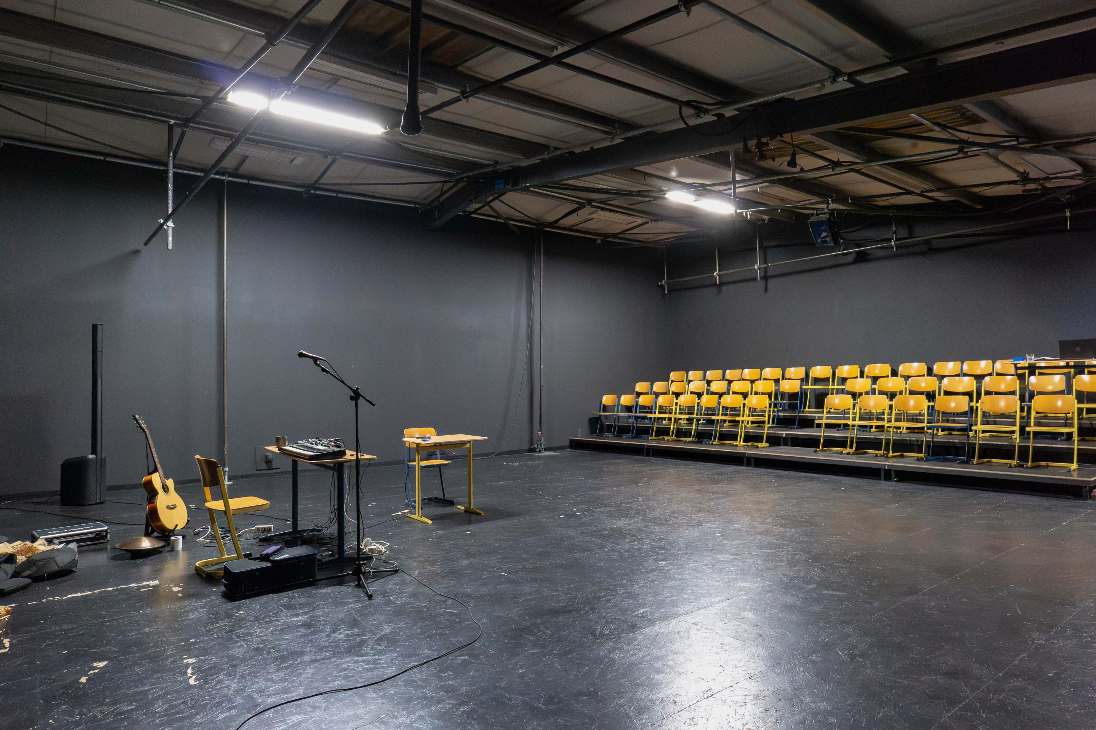The image shows a sparsely furnished performance space with a microphone, musical equipment, an acoustic guitar, a table, and rows of yellow chairs against a gray wall.