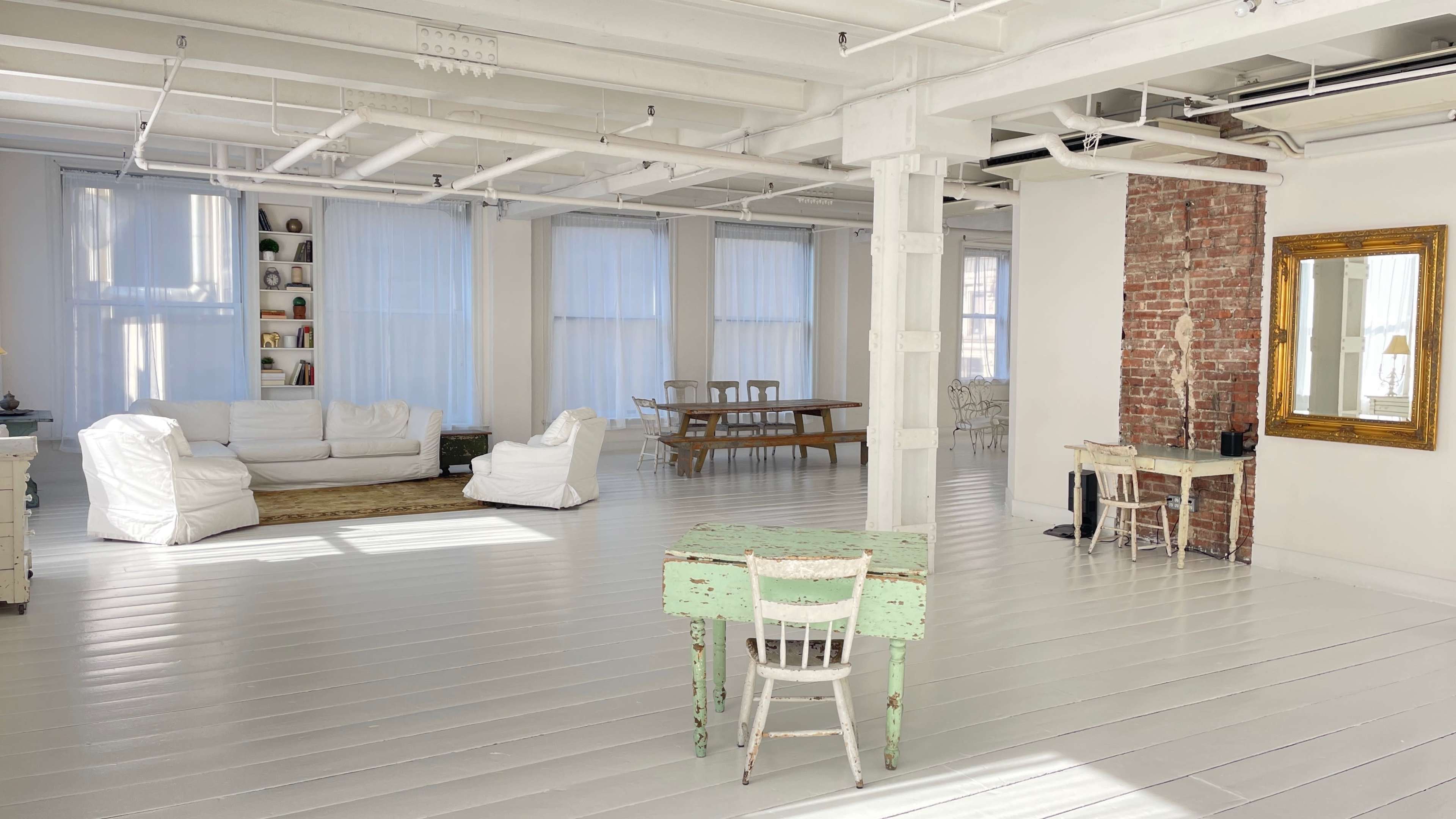A spacious, well-lit room with white wooden floors, furnished with various seating areas, a long dining table, and a green table with chairs.