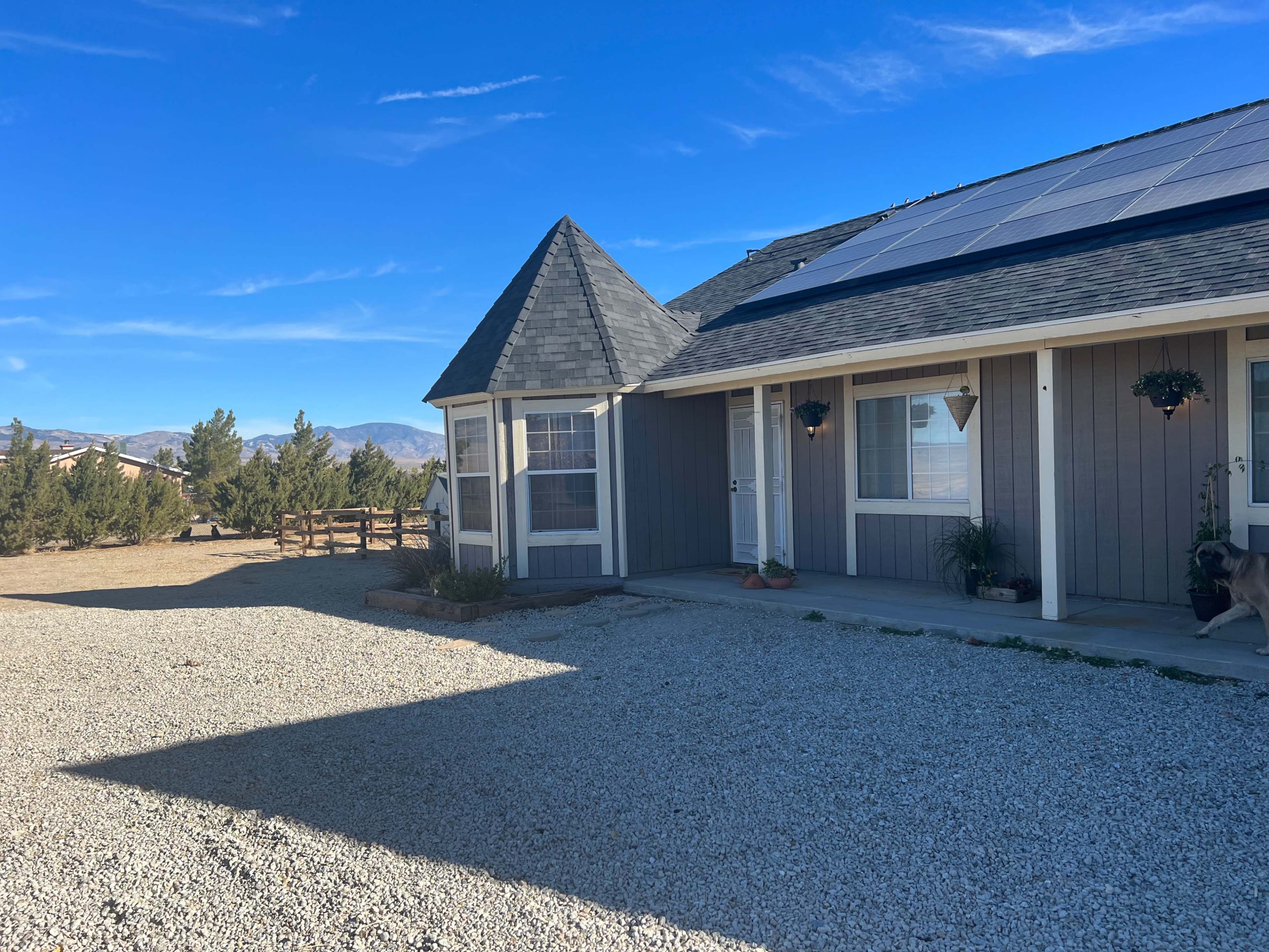 A house with a gray exterior and a peaked roof is situated next to a gravel driveway and solar panels, surrounded by sparse trees and mountains in the background.
