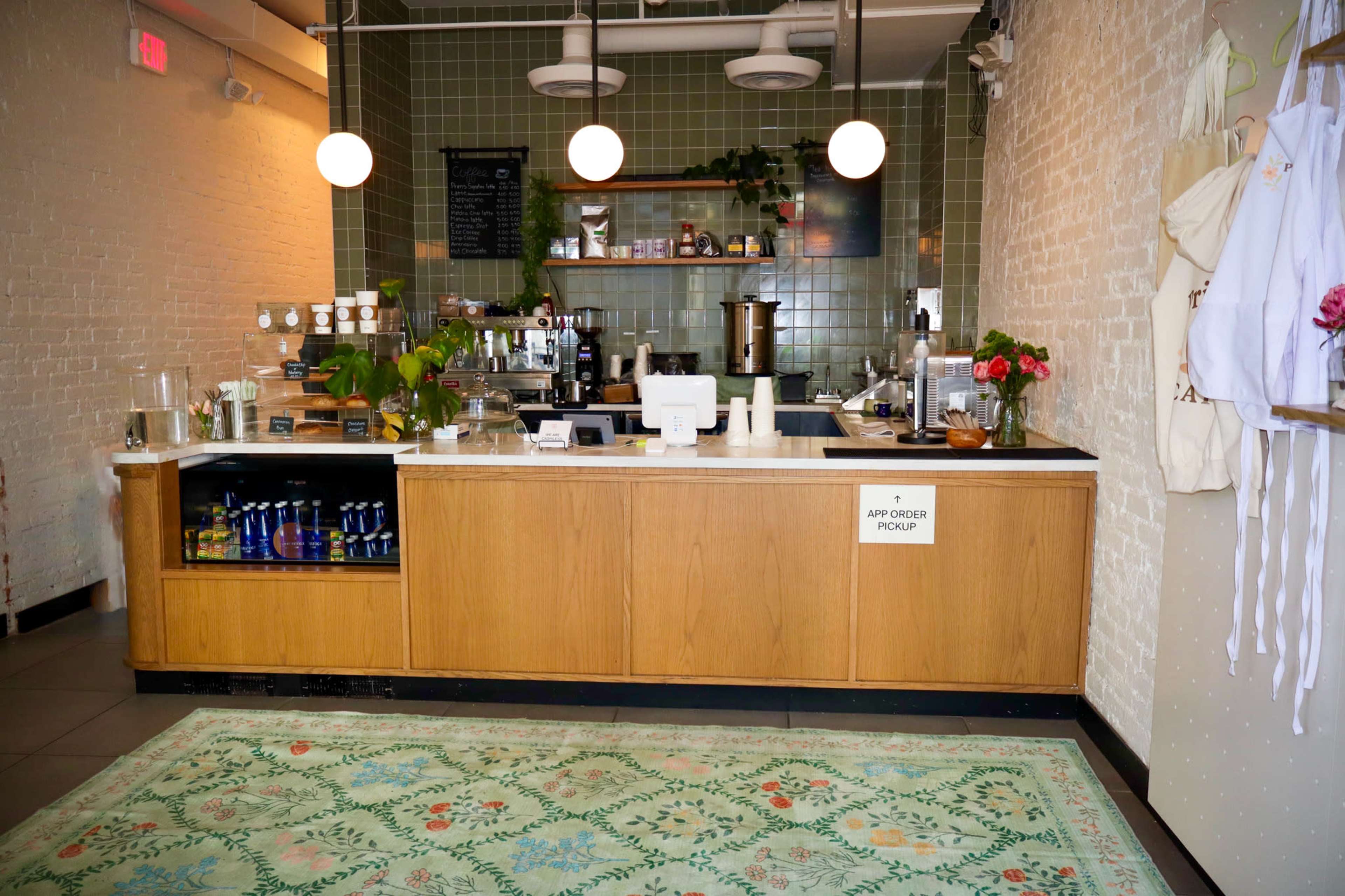 The image shows a café counter with a wooden front, a menu board above, various beverages on display, and a decorative green tile wall.