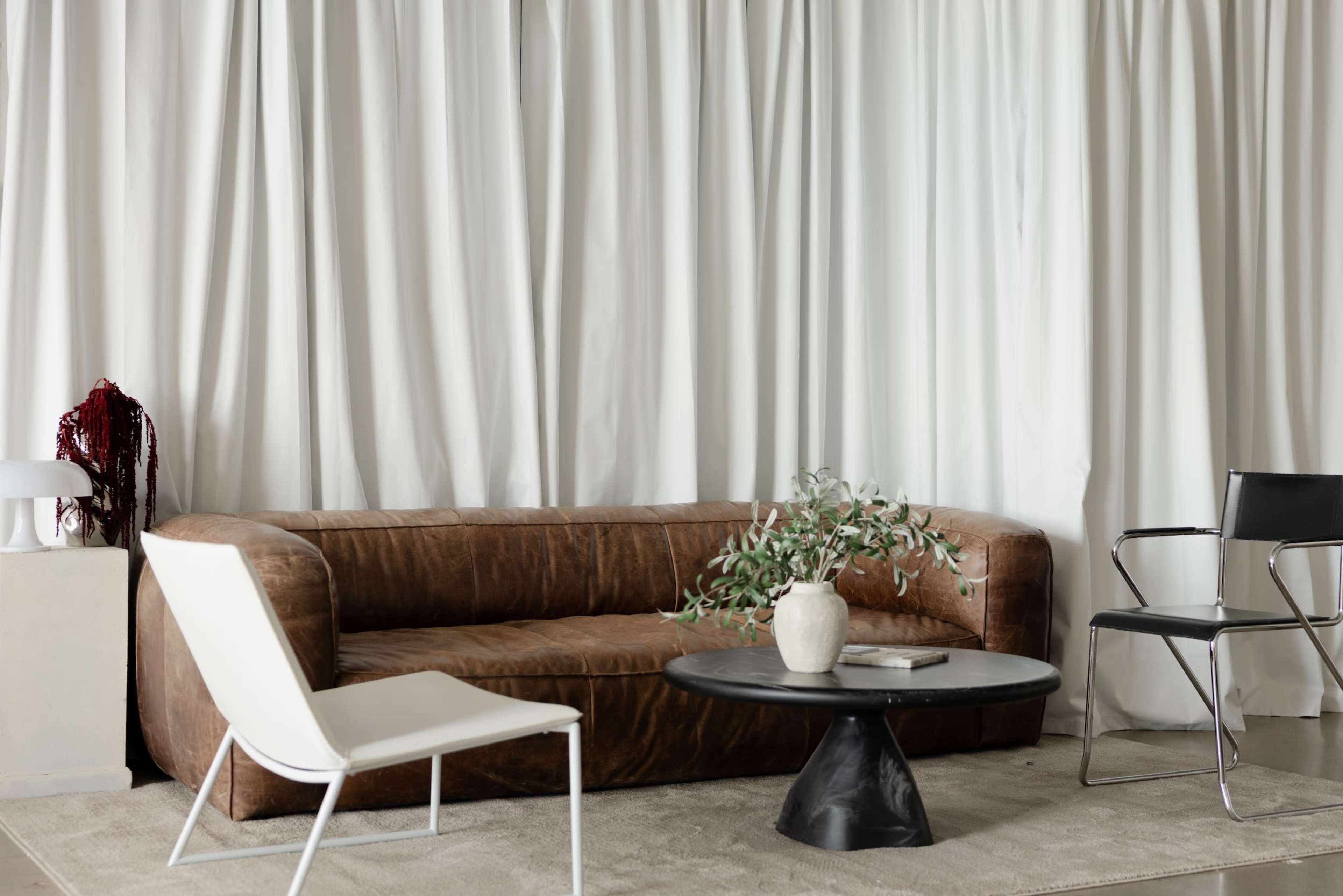 A brown leather sofa is positioned in a room with white draped curtains, accompanied by a black round table and a white chair.