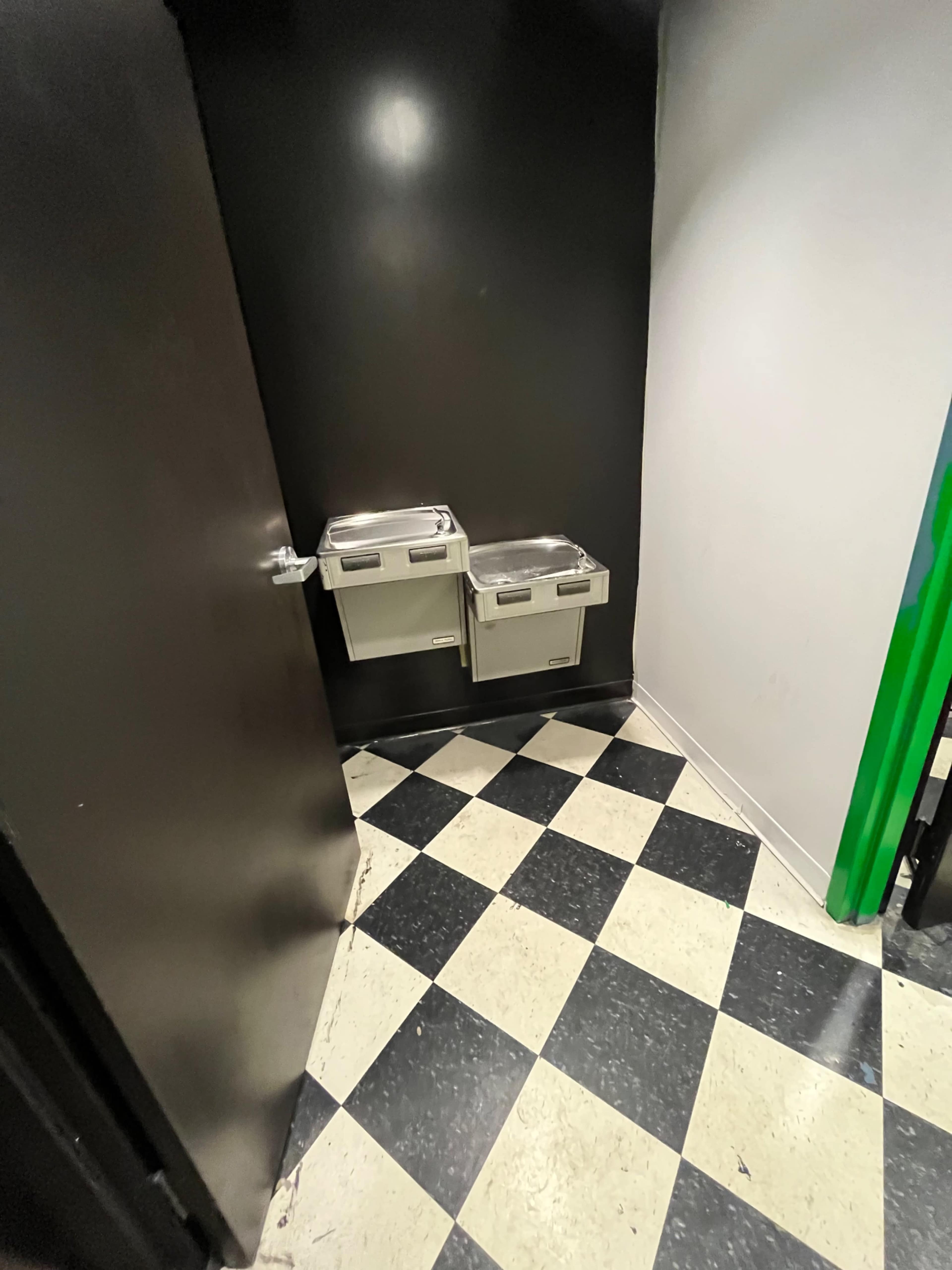 A corner of a room with two drinking fountains mounted on a black wall, adjacent to a door, and a checkered floor in black and white.