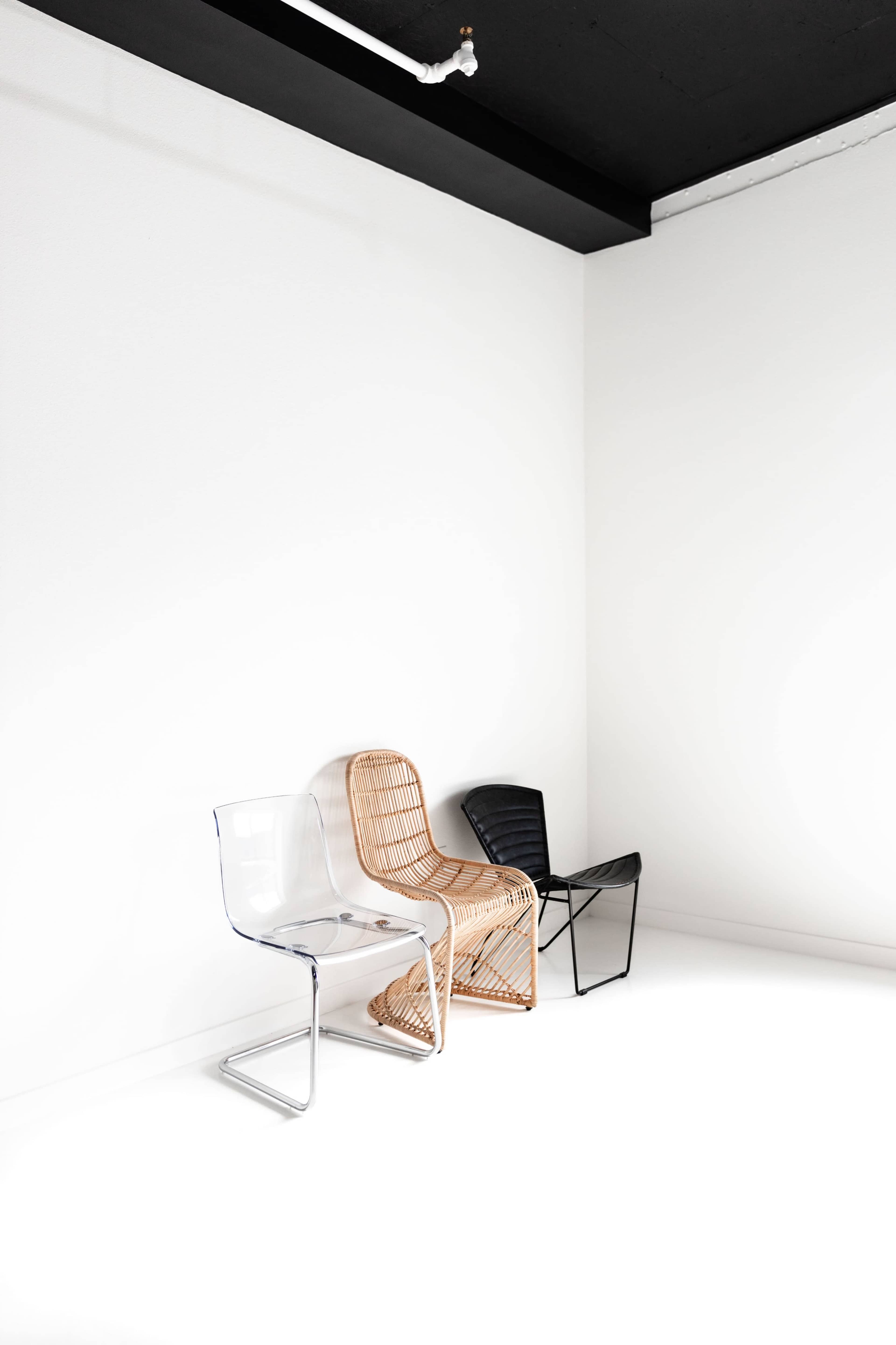 Three chairs in a minimalist space are positioned against a white wall, with one made of clear material, one in a natural wicker finish, and another in black metal.