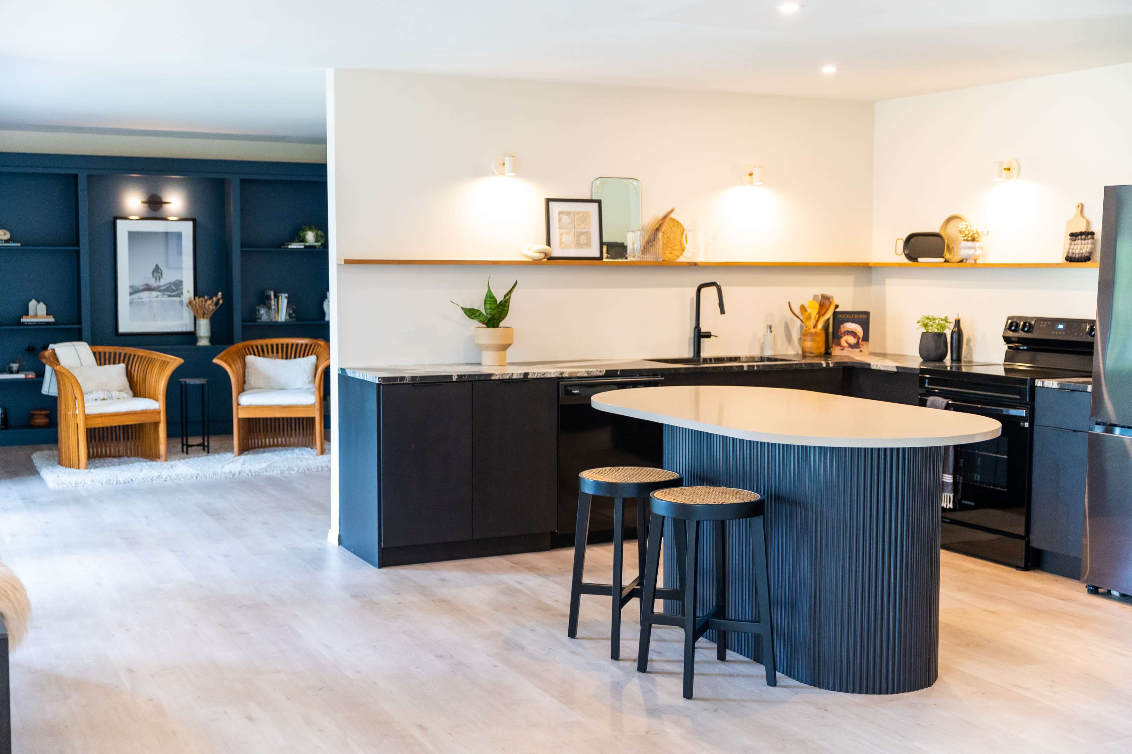 The image shows a modern kitchen with a central island, black cabinetry, and a seating area featuring two rattan chairs in the background.