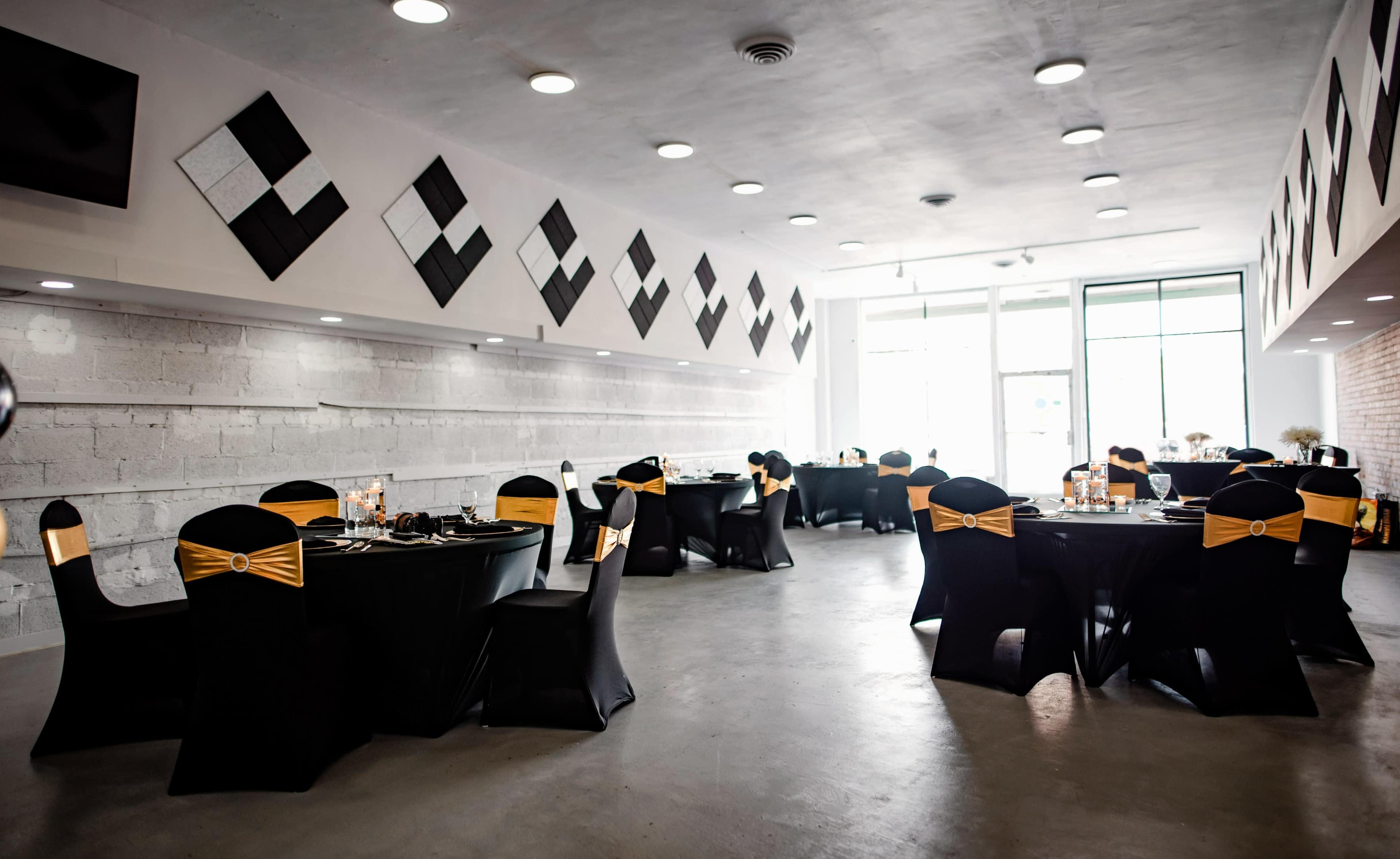 A modern event space set up with black tables and chairs adorned with gold sashes, and minimalistic decor on the walls.