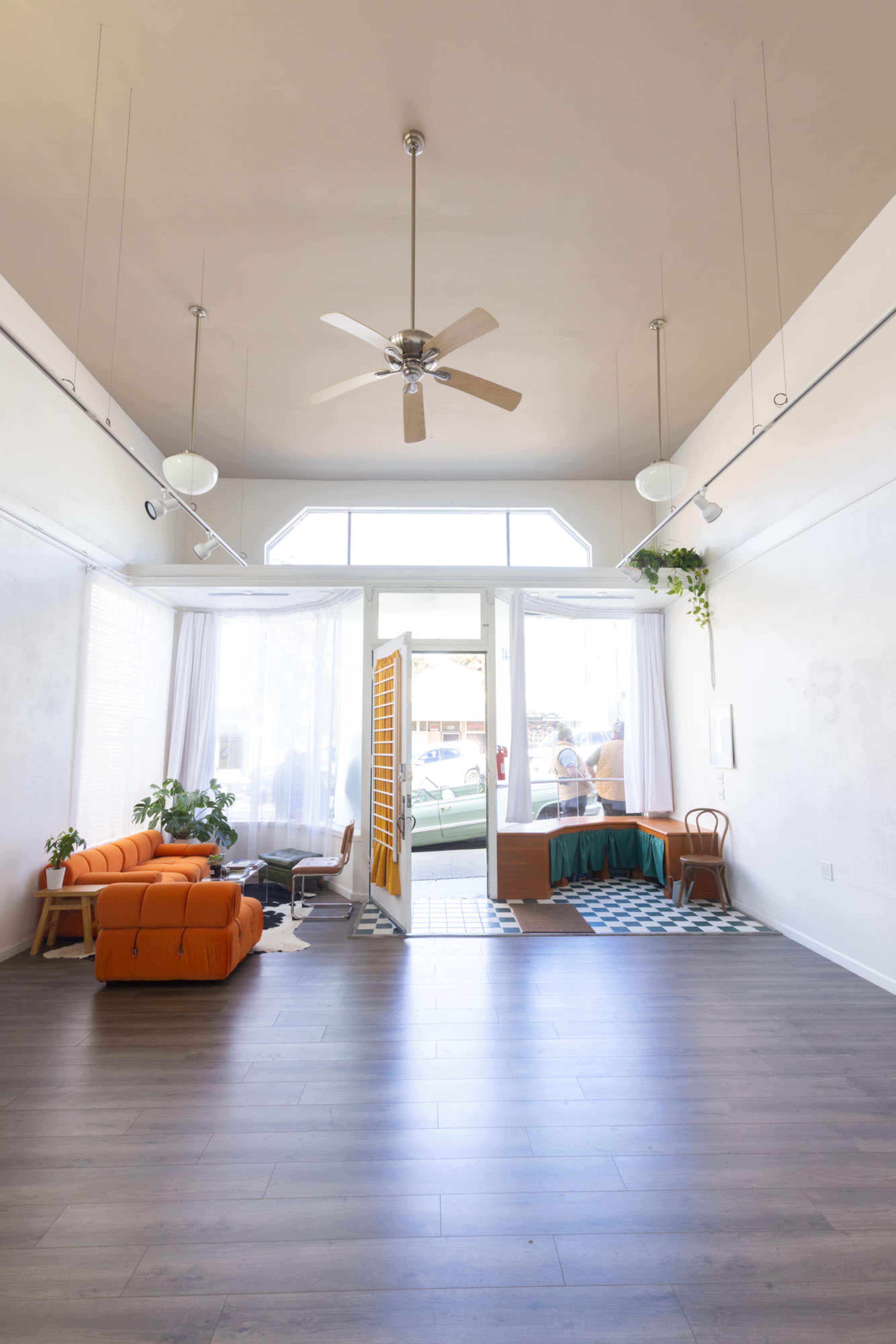 The image shows a spacious room with a large glass entrance, an orange sofa, a small table, and potted plants.