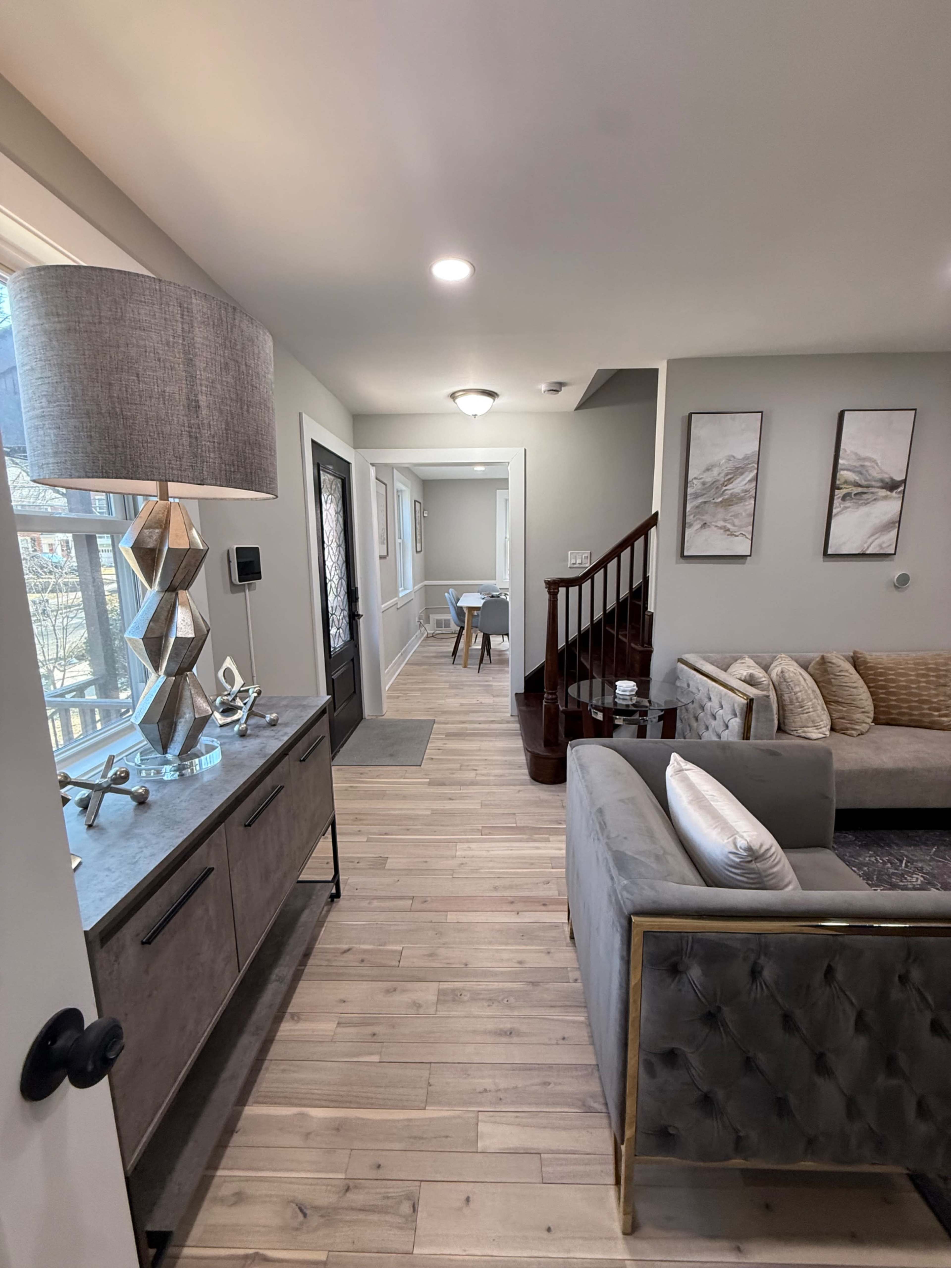 The image shows a modern living room with a gray sofa, a decorative lamp on a side table, and a staircase leading to another area of the house.