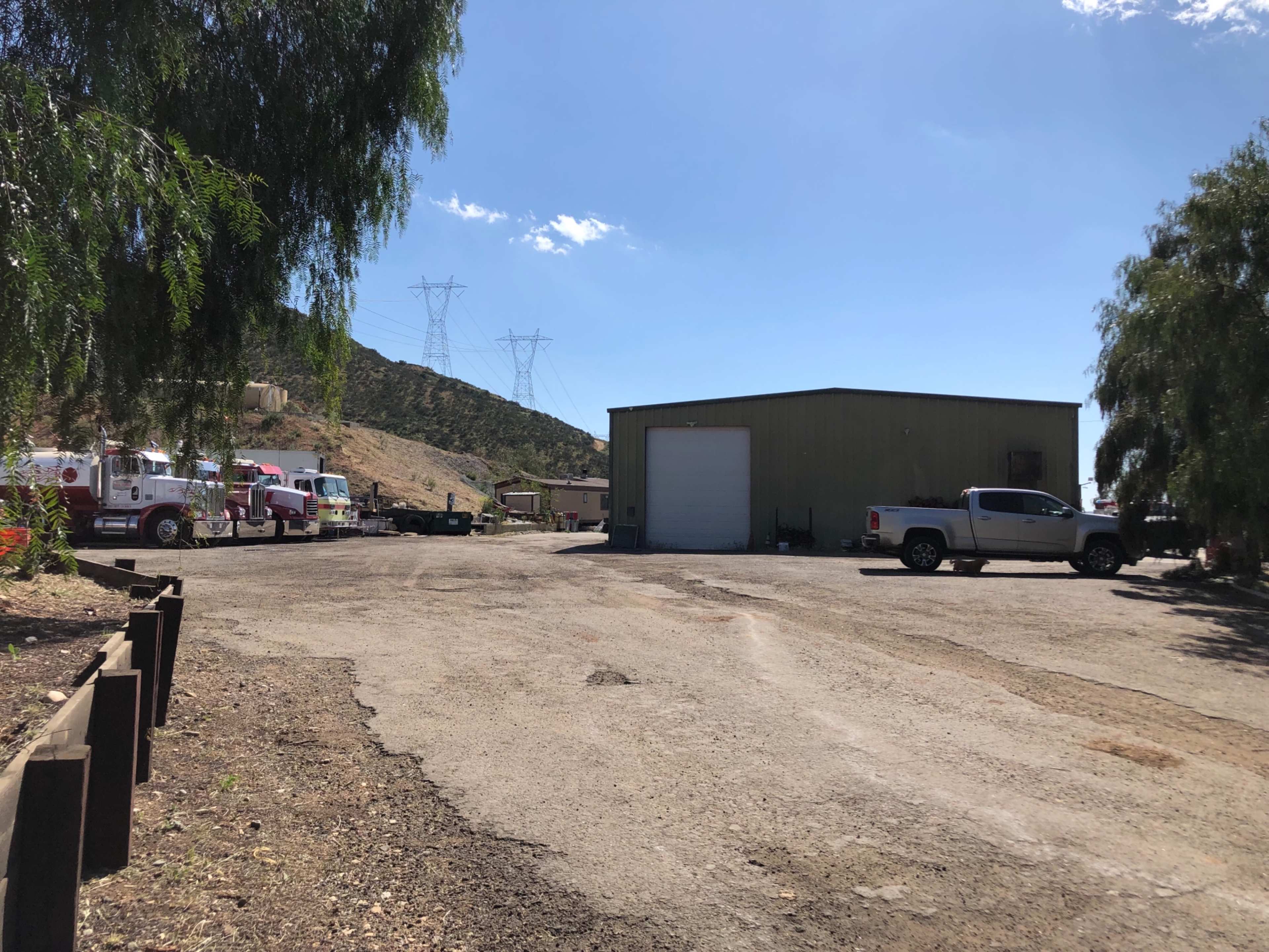 A dirt parking lot features a metal building, several vehicles including a fire truck, and power lines in the background.