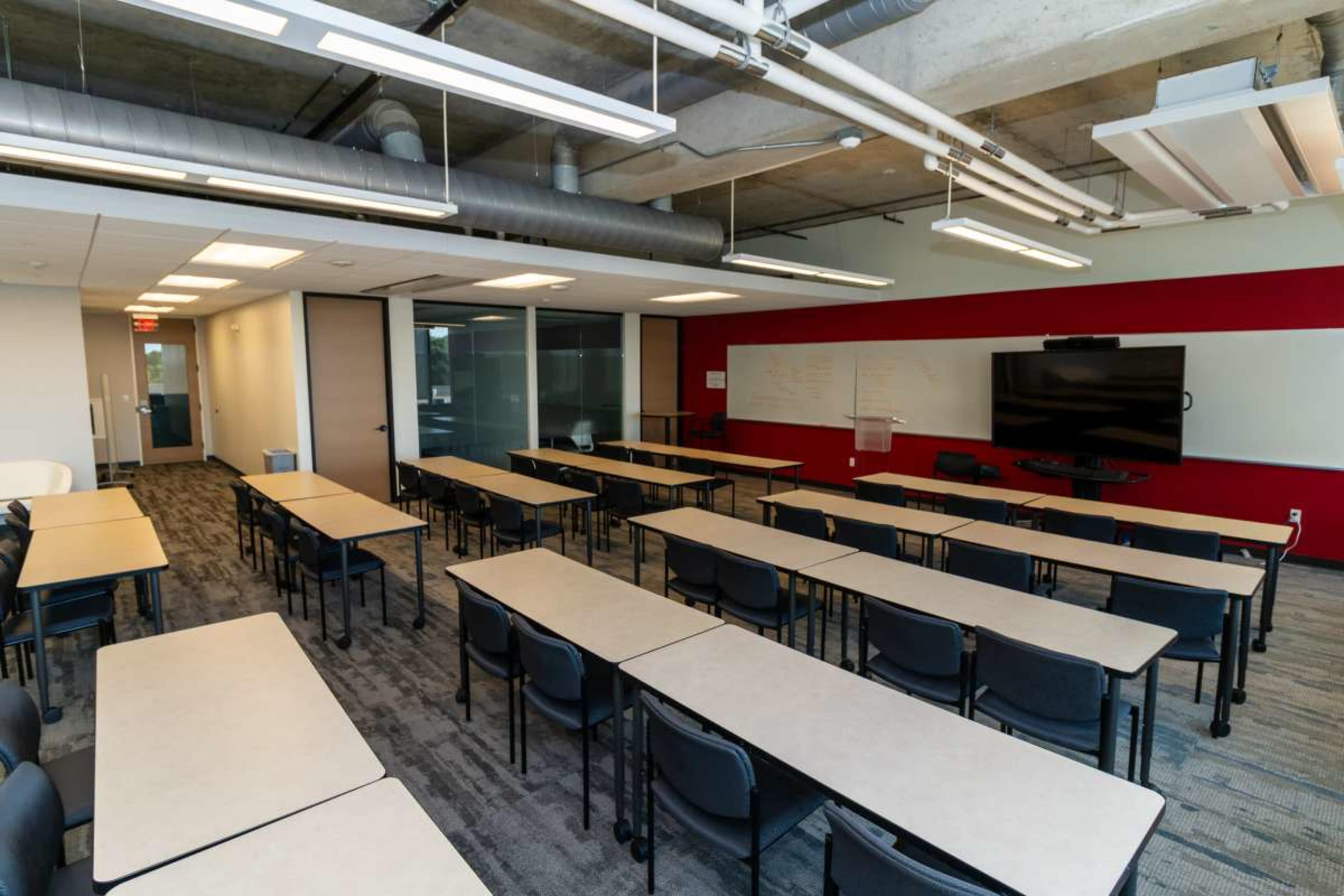 The image shows a modern classroom with rows of tables and chairs, a large screen at the front, and a whiteboard on a red wall.