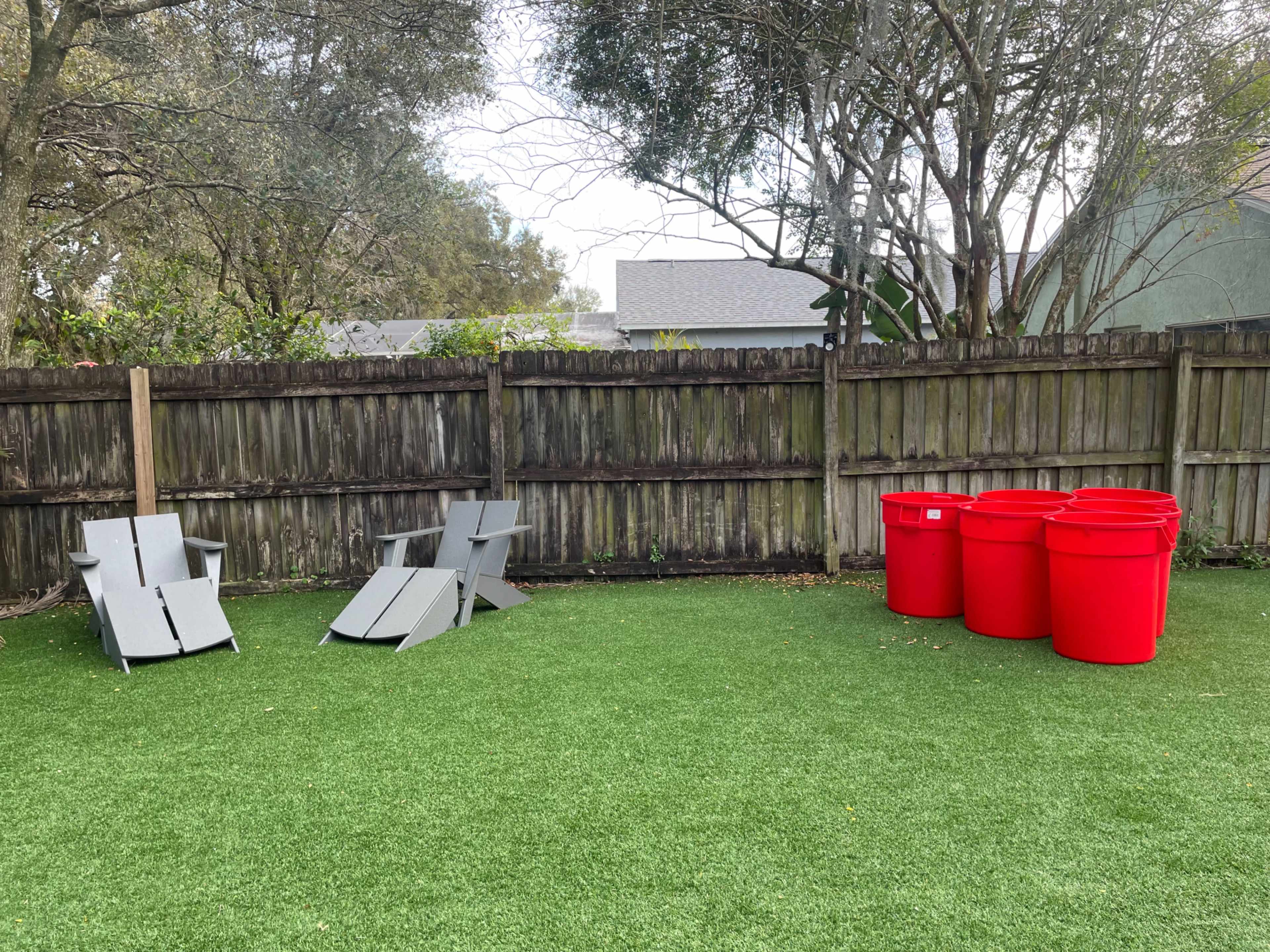 The image shows a backyard with artificial turf, two gray folding chairs near a wooden fence, and three red trash bins arranged together.
