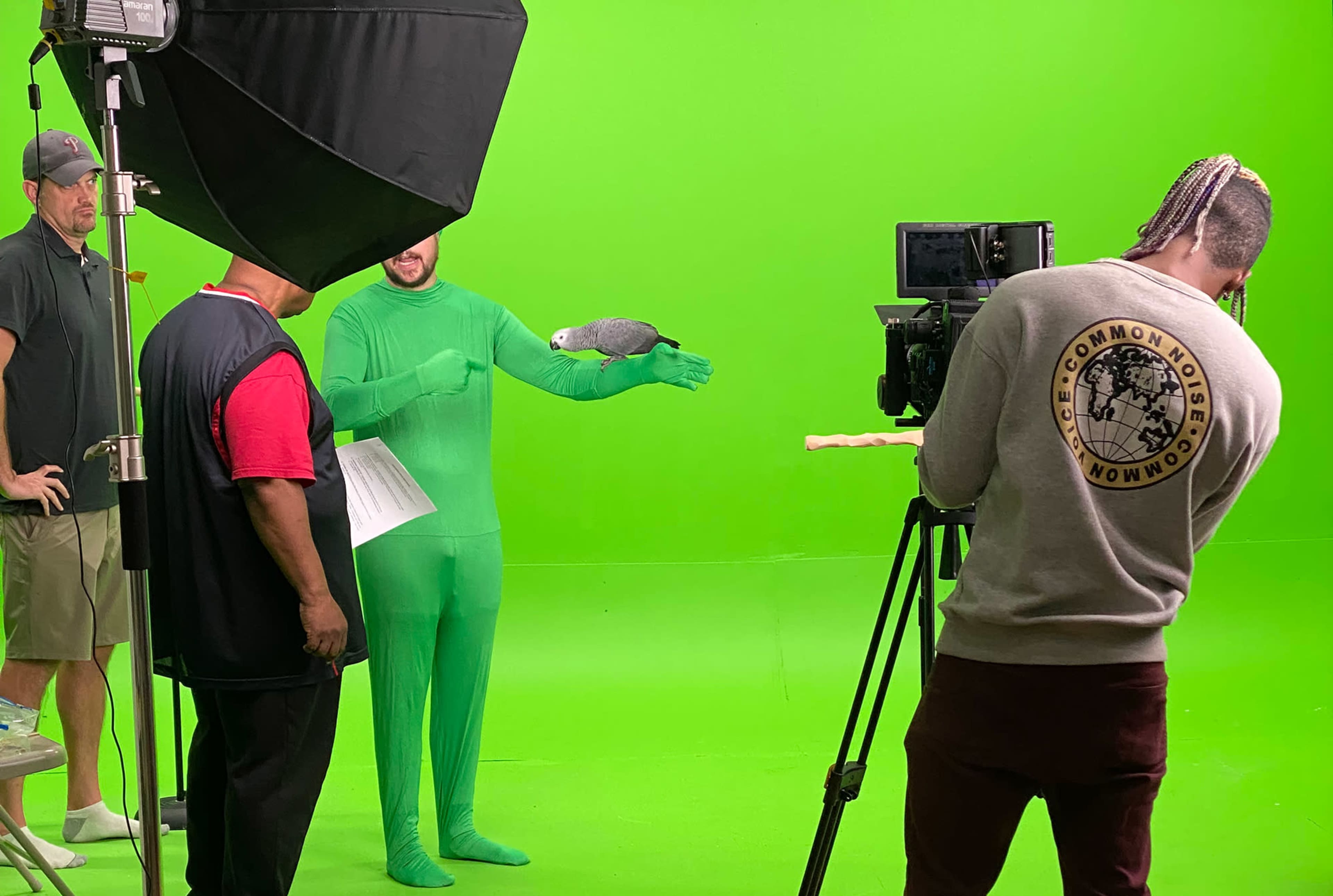 A group of people is filming a scene with a man in a green suit holding a bird in front of a green screen.