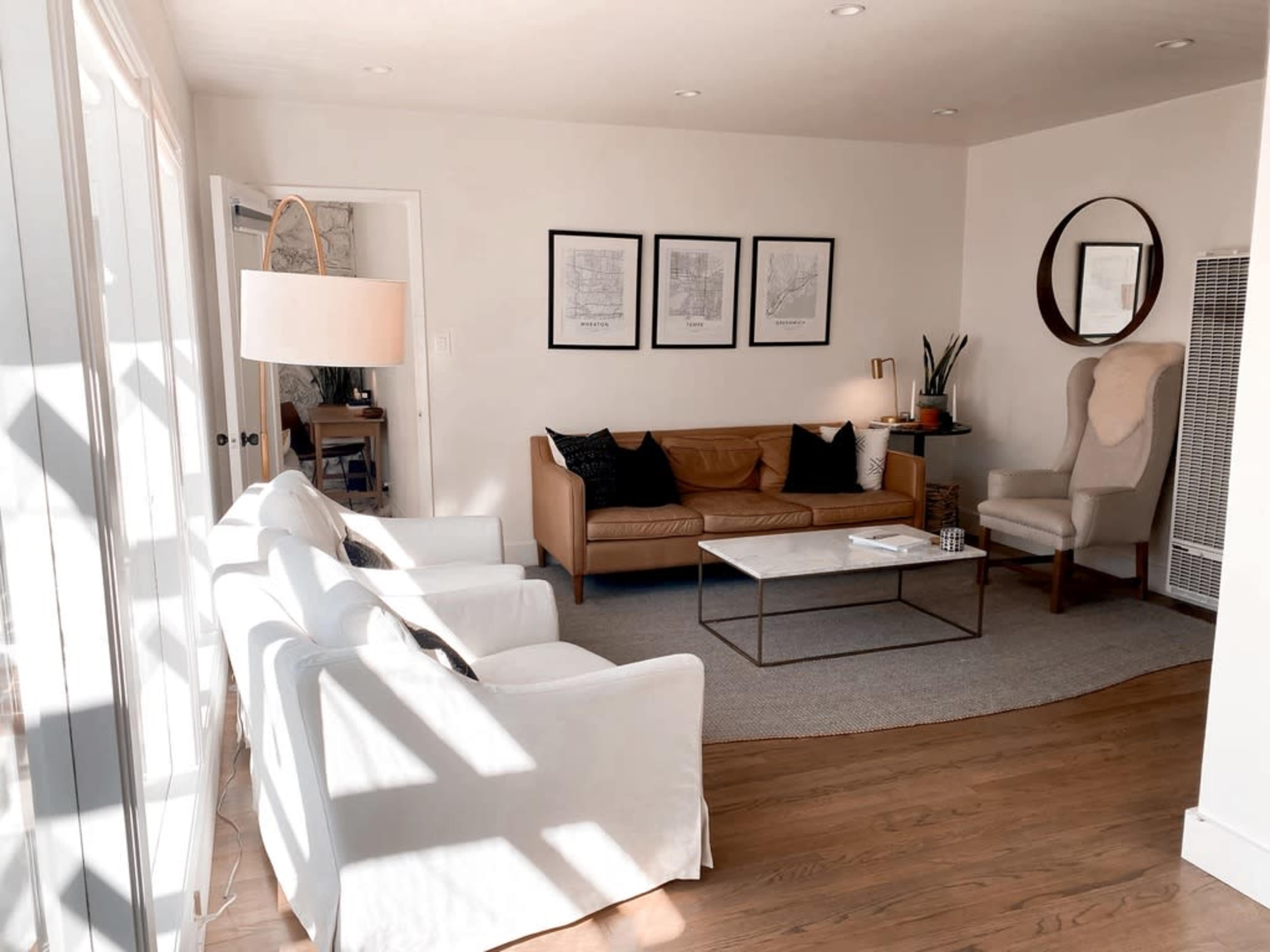 A bright living room featuring a tan sofa, two white chairs, a coffee table, and framed artwork on the walls.