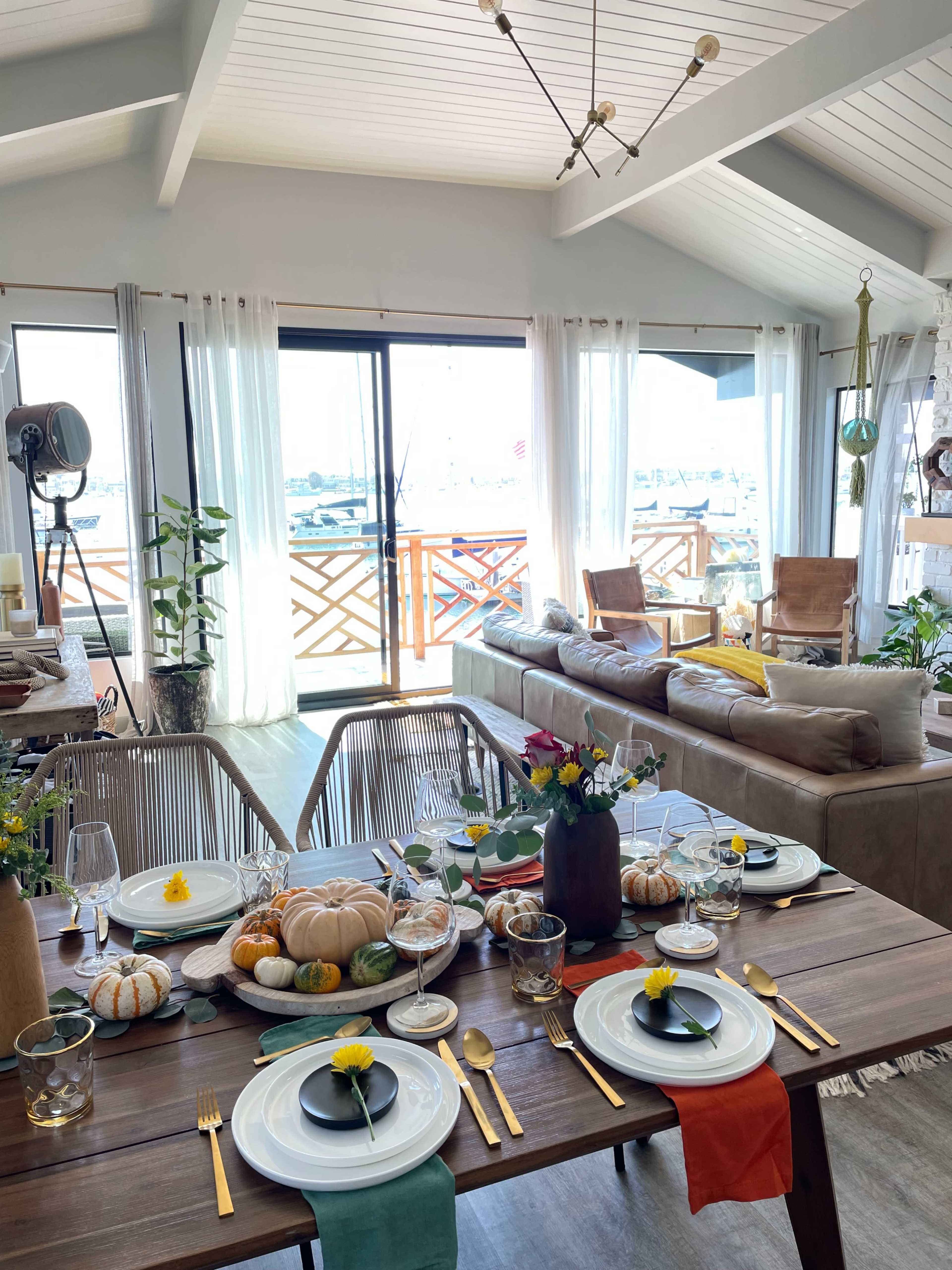 A dining table set with colorful pumpkins and flowers is displayed in a sunlit living space overlooking a waterfront balcony.