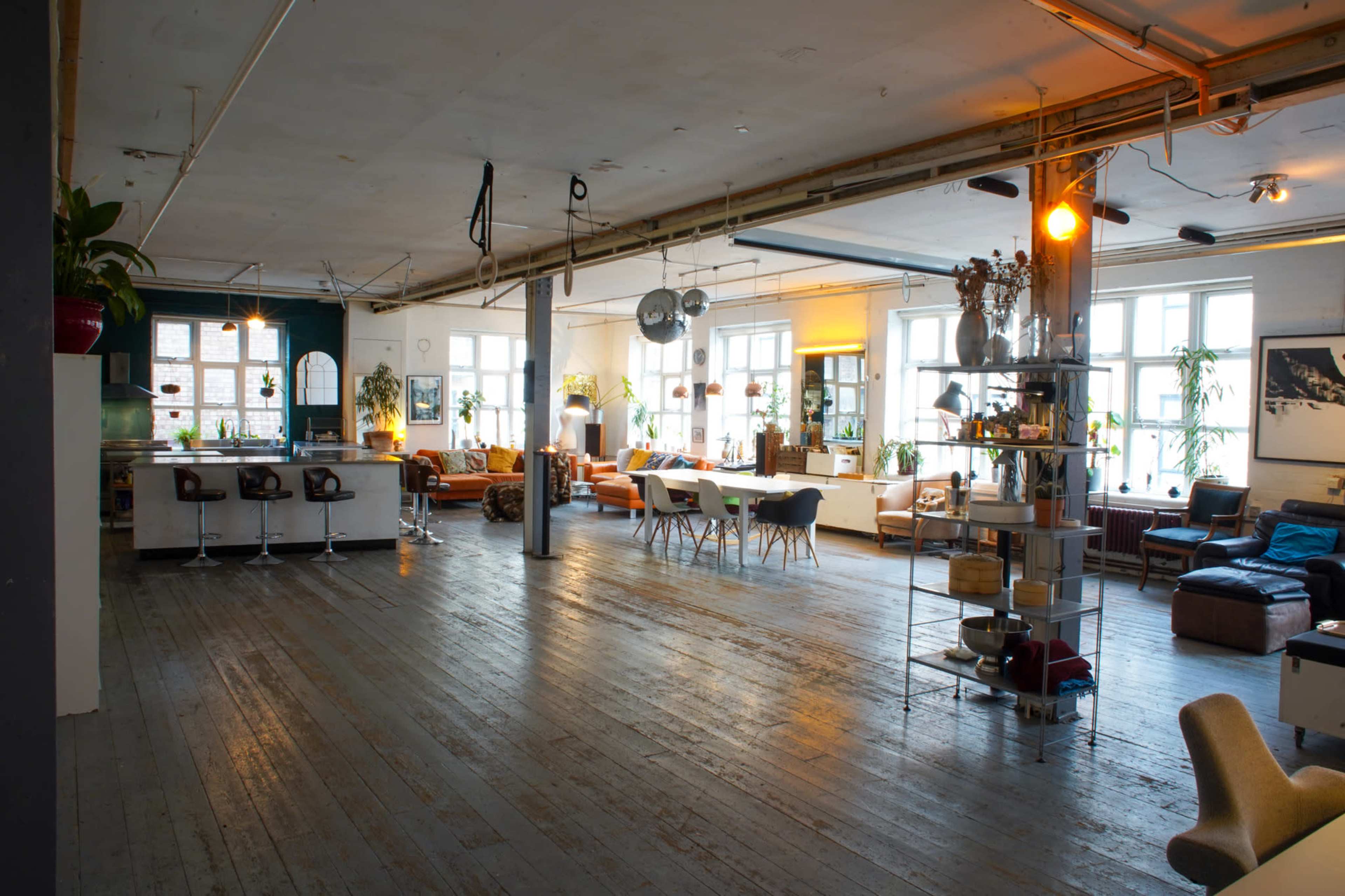The image shows a spacious, airy loft with wooden floors, large windows, and a mix of seating areas featuring sofas and chairs, along with a dining table and bar stools.