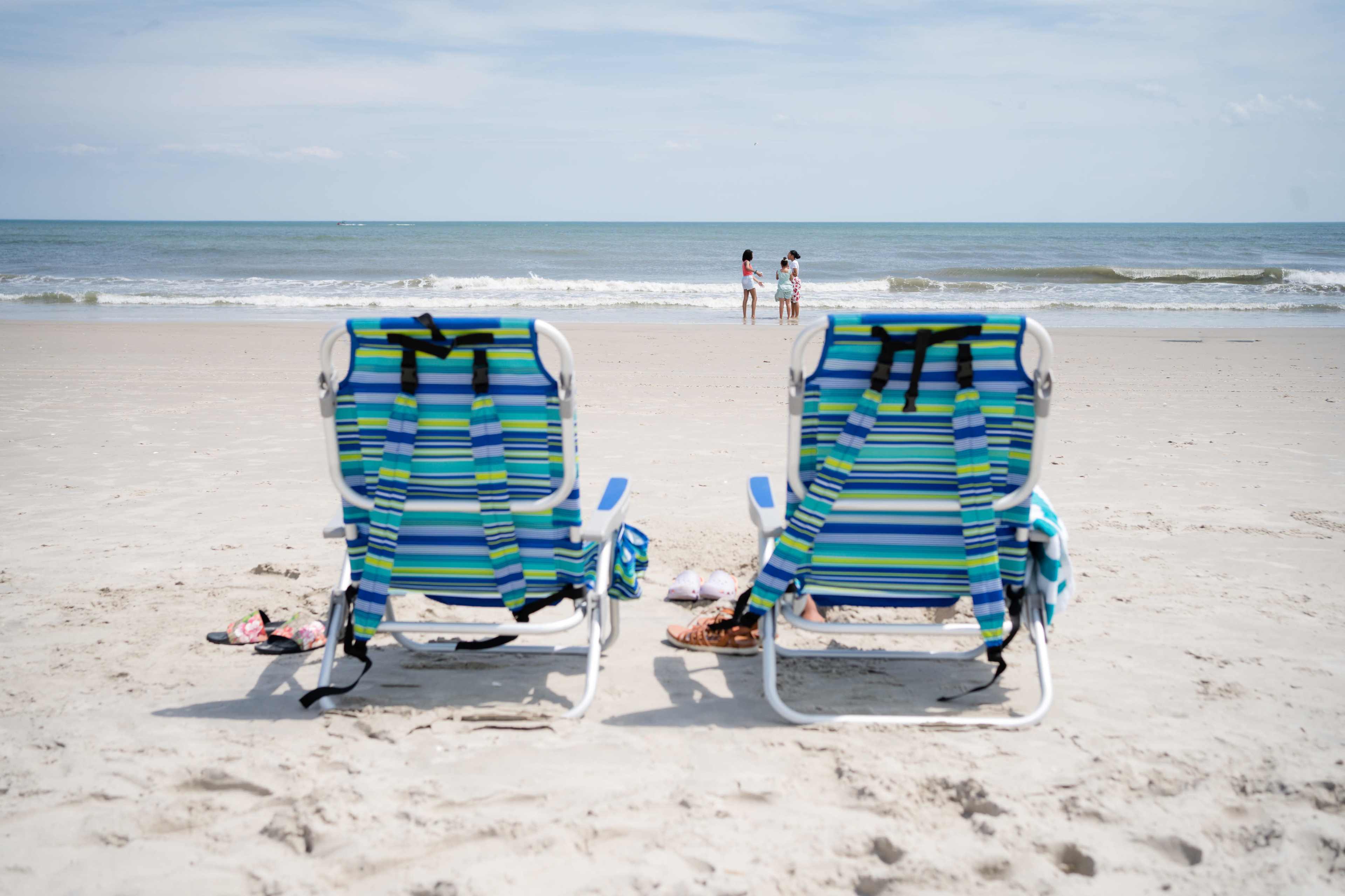Two blue and green striped beach chairs are positioned on a sandy shore, facing the ocean where two people are standing in the water.