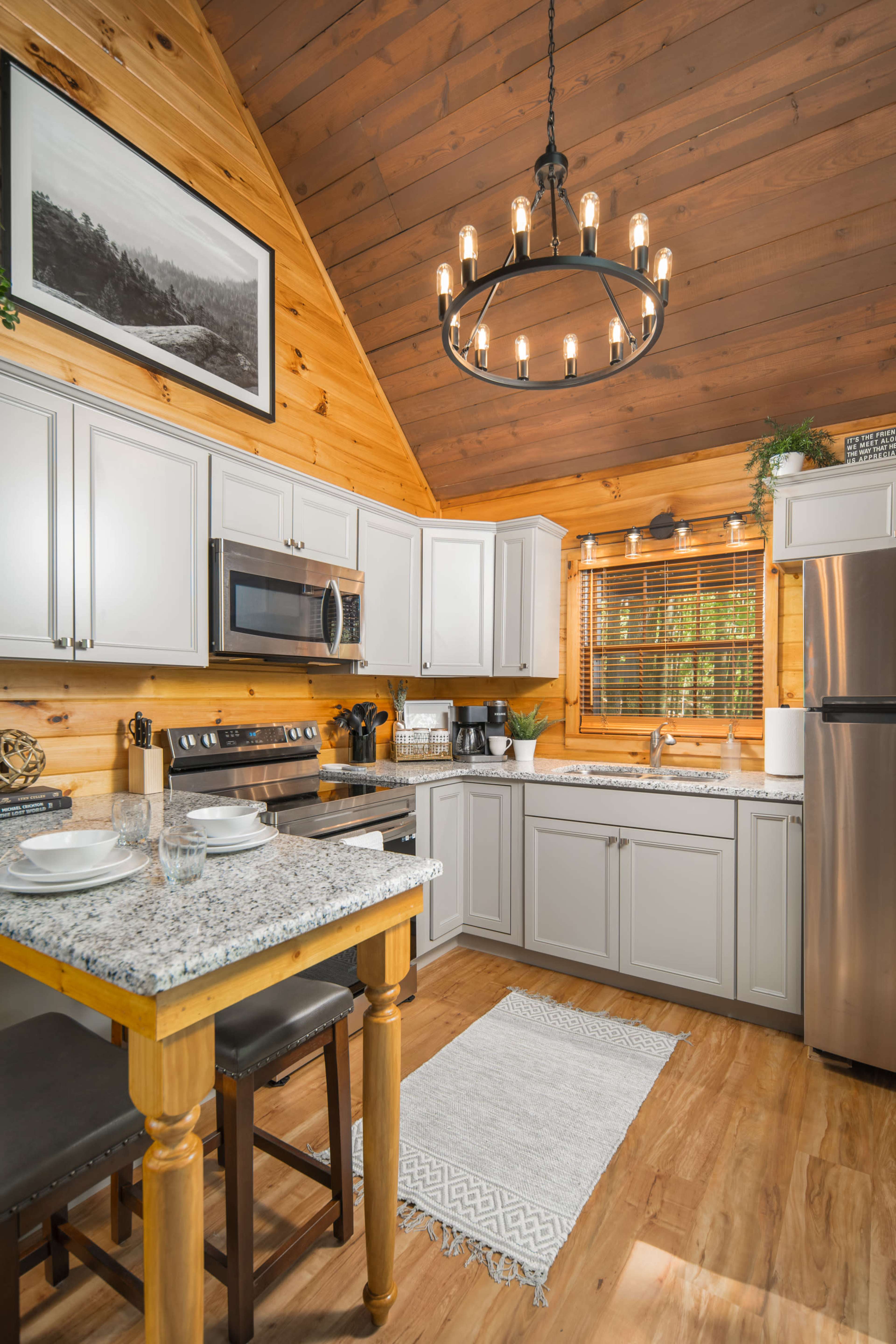 A modern kitchen with light gray cabinets, granite countertops, a stainless steel refrigerator, and wooden accents, all set under a chandelier in a cabin-style interior.