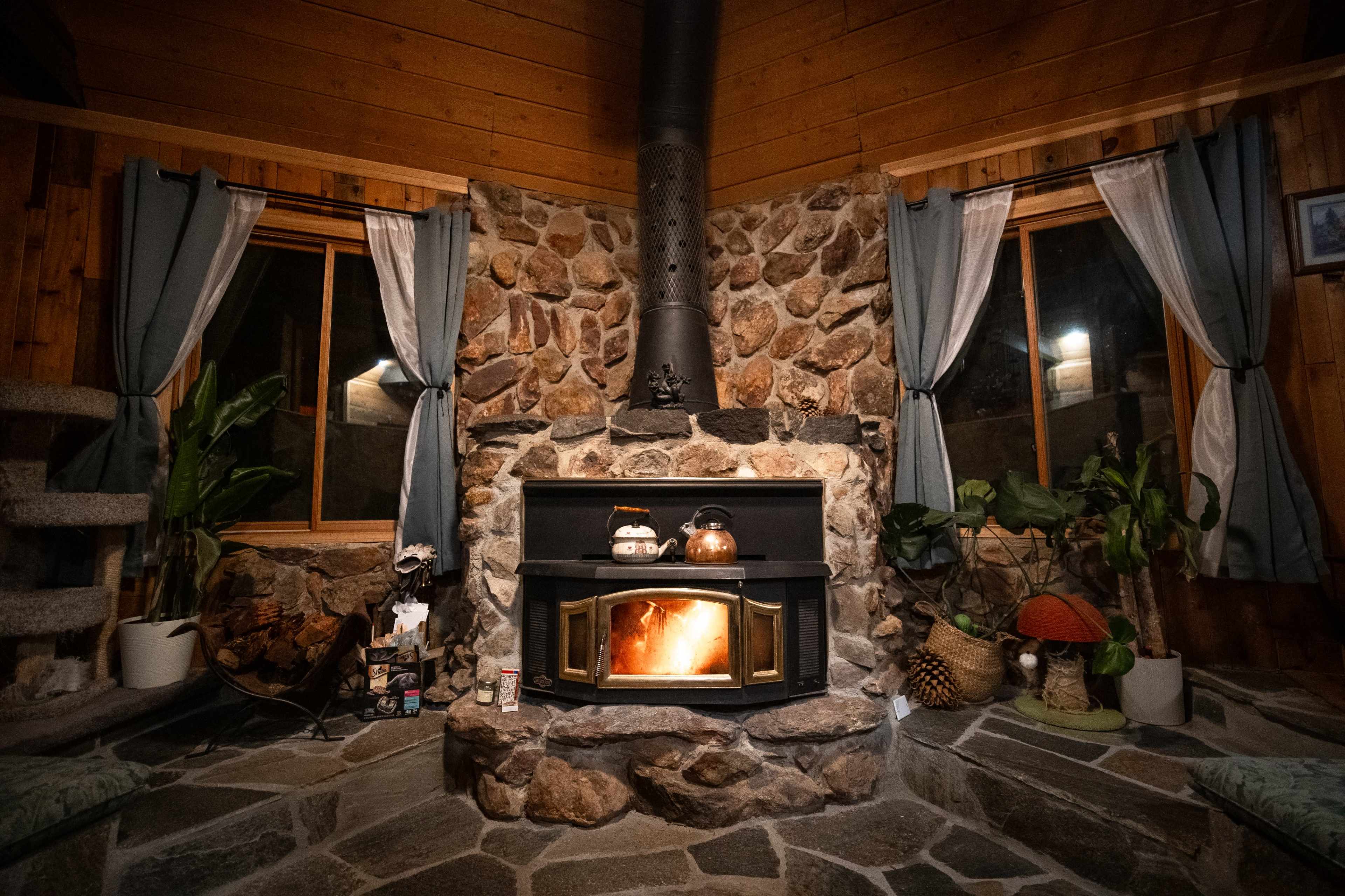 A stone fireplace with a wood-burning stove is centered in a cozy room with wooden walls and large windows.