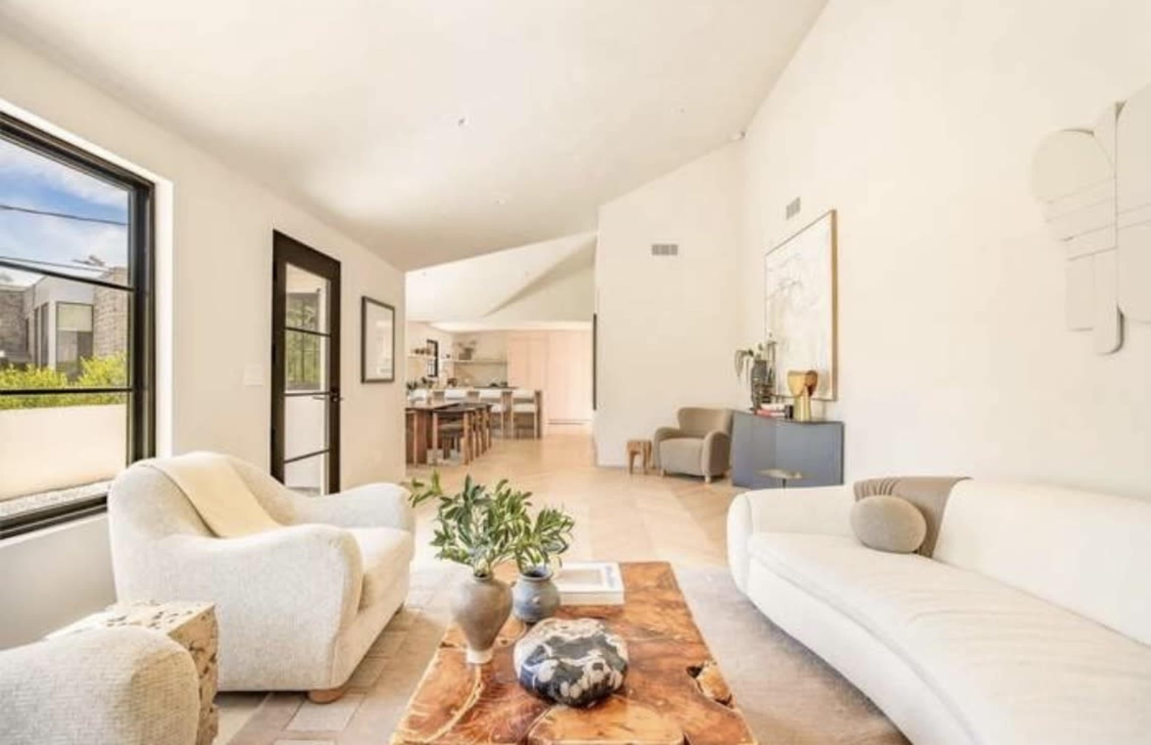 The image shows a light-filled living room featuring a white sofa and armchair, a wooden coffee table, and a dining area visible in the background.