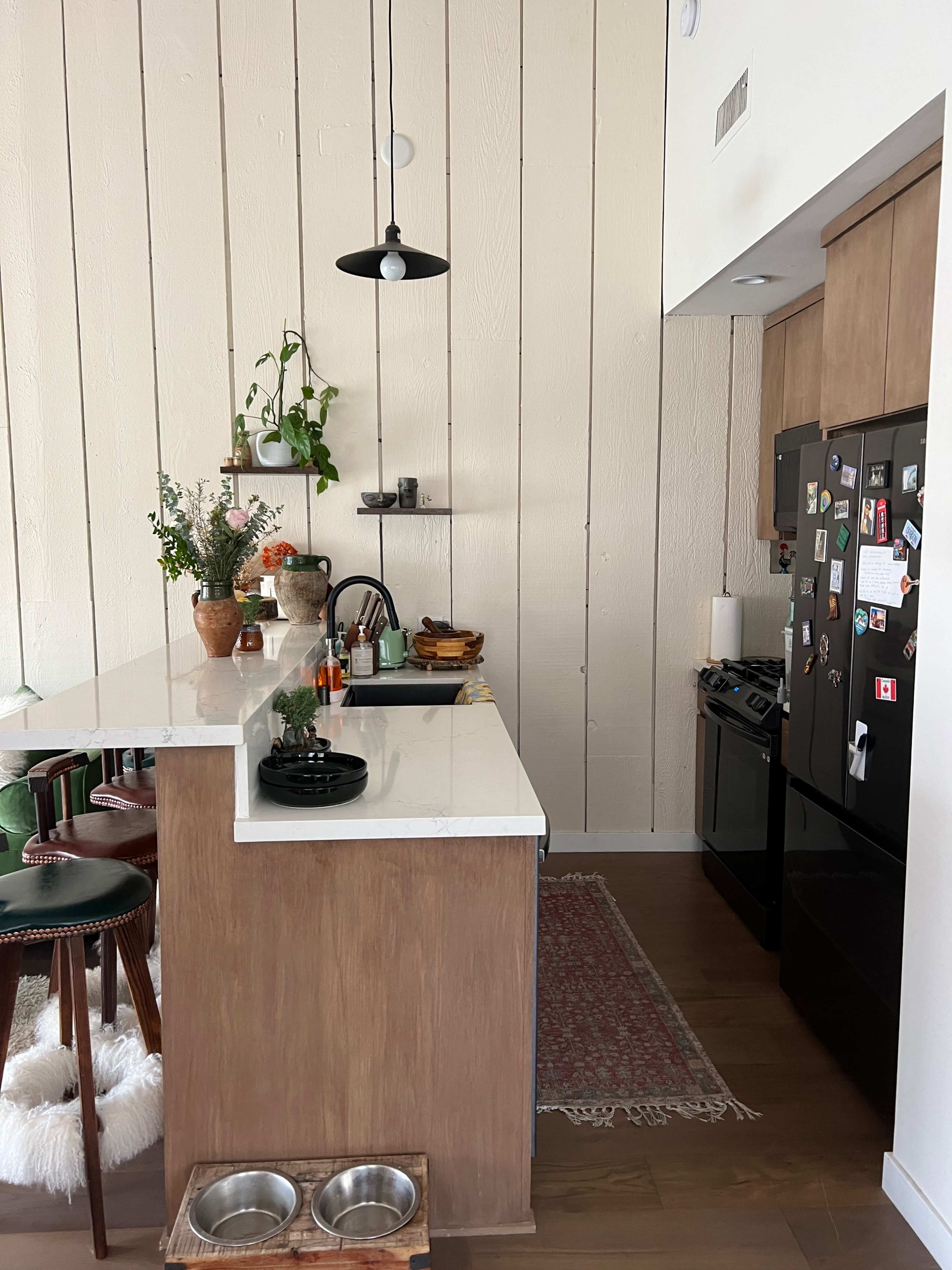 The image shows a modern kitchen with a light-colored wood panel wall, a countertop with bar seating, and various kitchen appliances and decor.