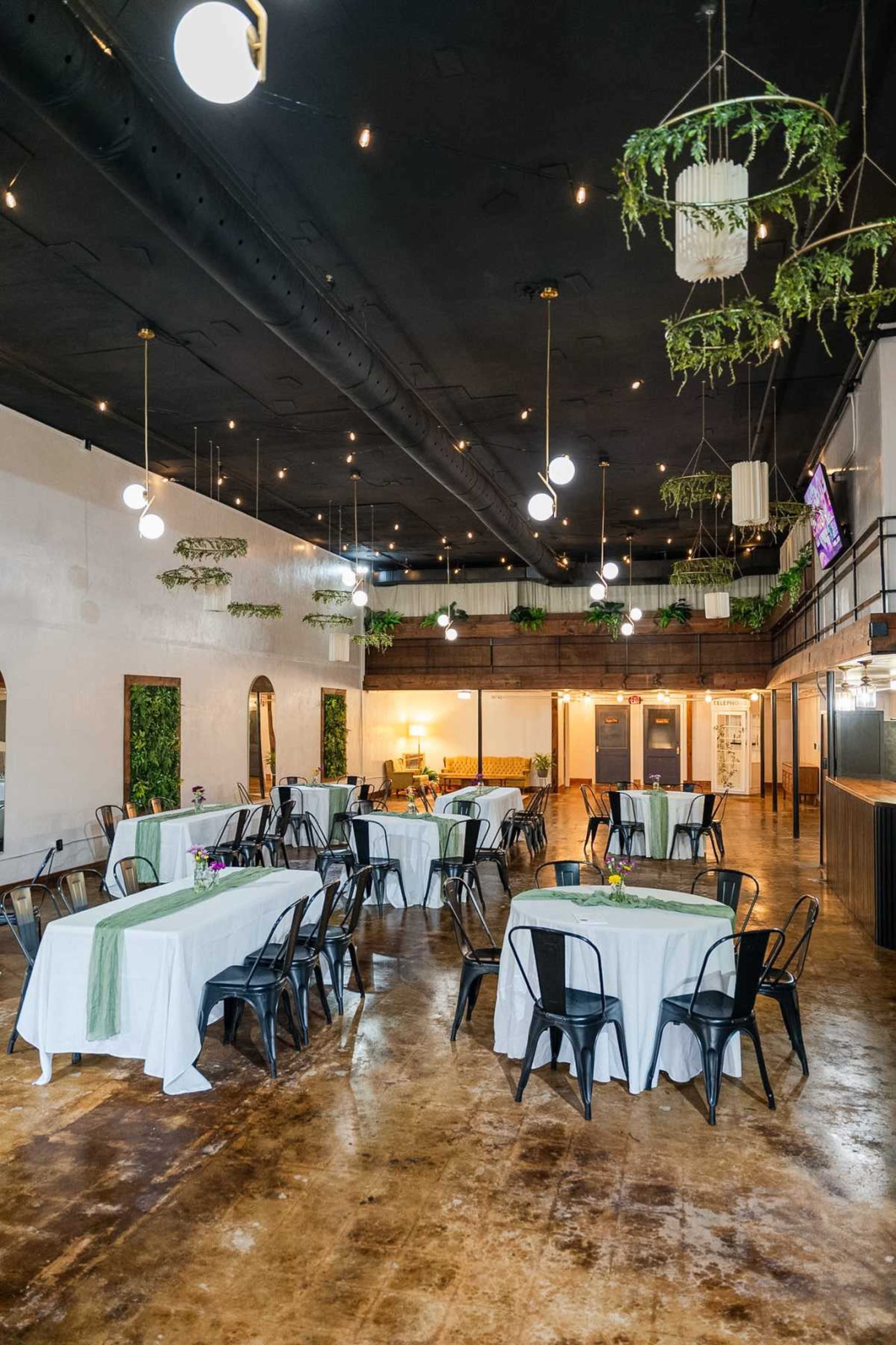 The image shows a spacious restaurant interior with tables set for dining, featuring white tablecloths and black metal chairs, illuminated by hanging light fixtures.