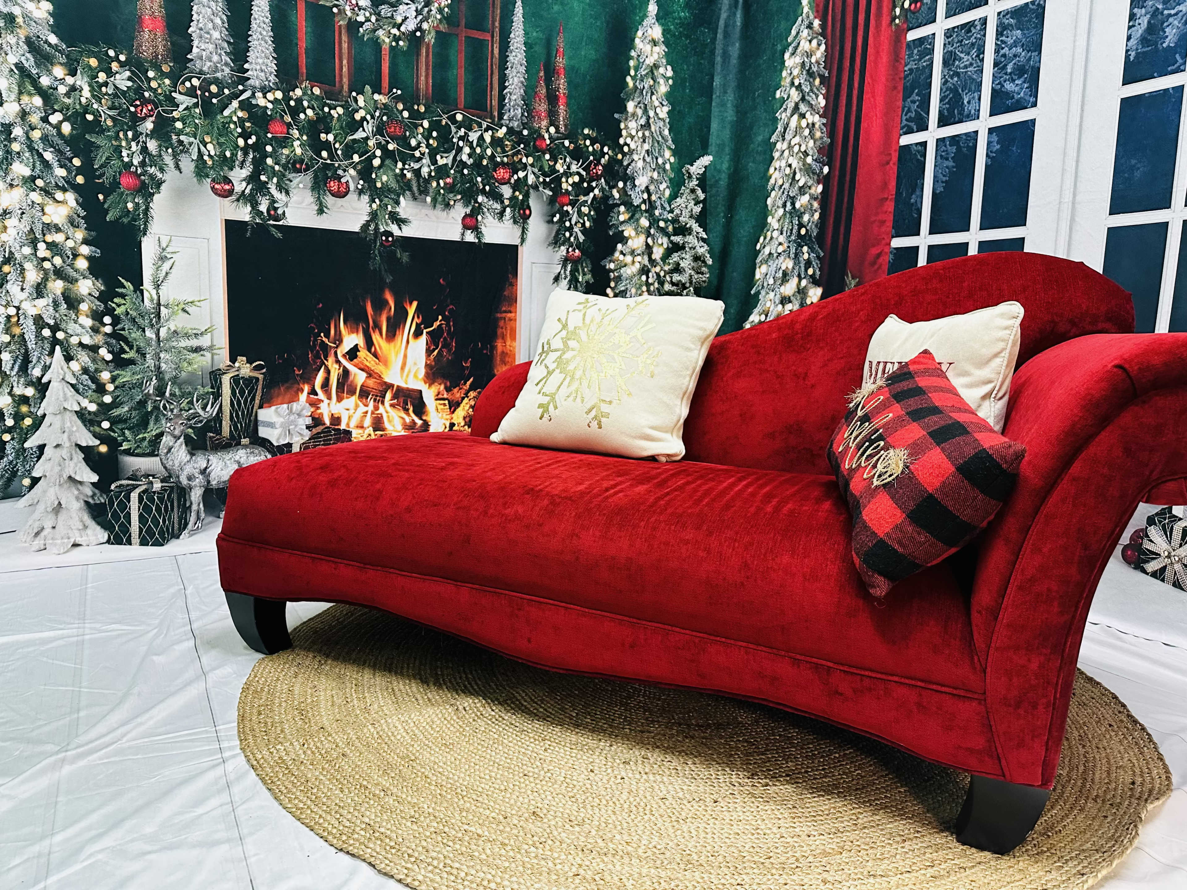 A red chaise lounge with decorative cushions is positioned in front of a festive backdrop featuring a fireplace and holiday-themed decorations.