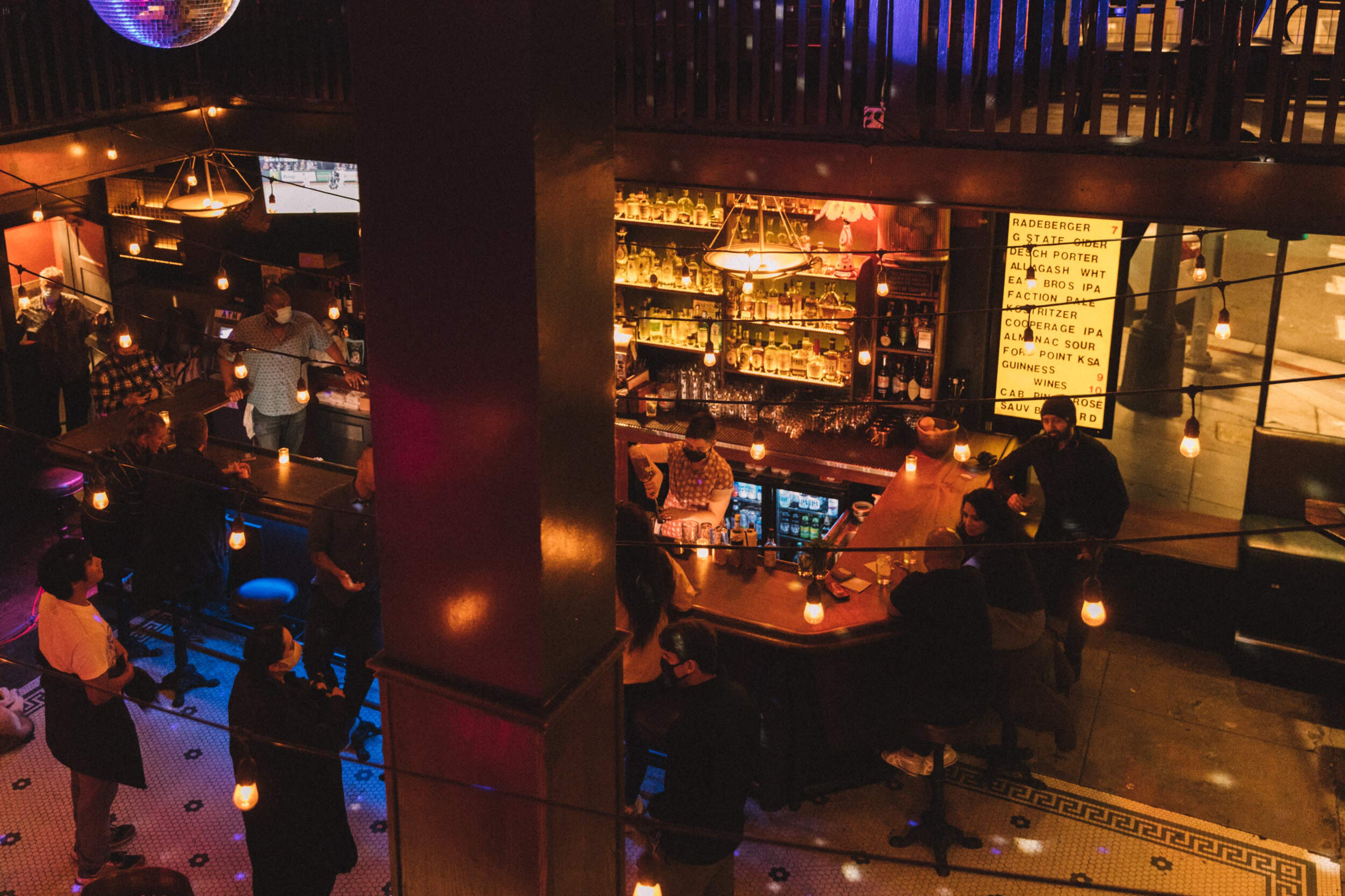 A lively bar scene features patrons gathered around a central counter with bottles displayed in the background, illuminated by dim lighting and hanging bulbs.