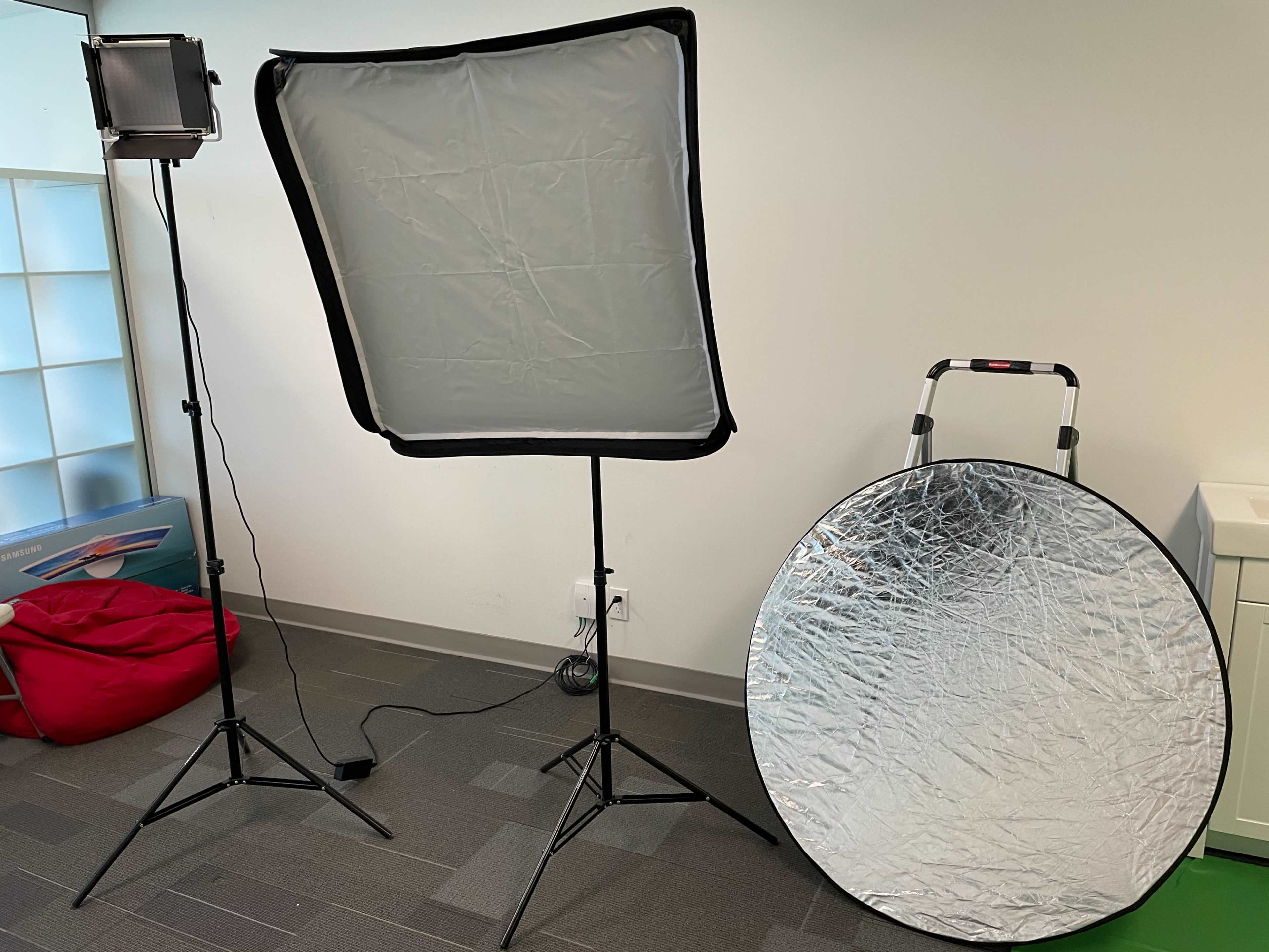A photography setup with a softbox light on a stand, a large circular reflector, and a portable light stand in an indoor workspace.