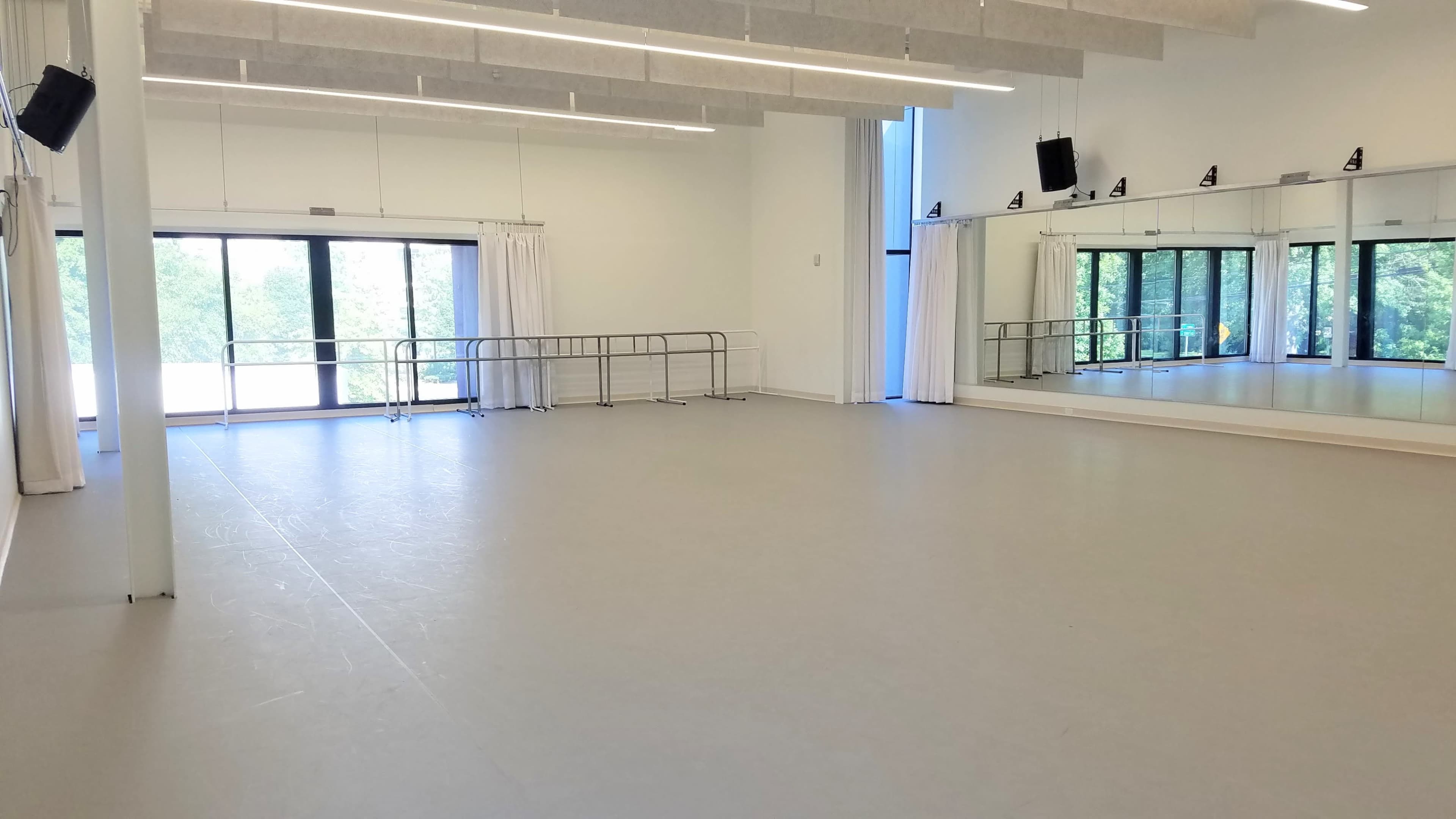 The image shows a spacious dance studio with large windows, mirrors on one wall, and ballet barres along the opposite side.