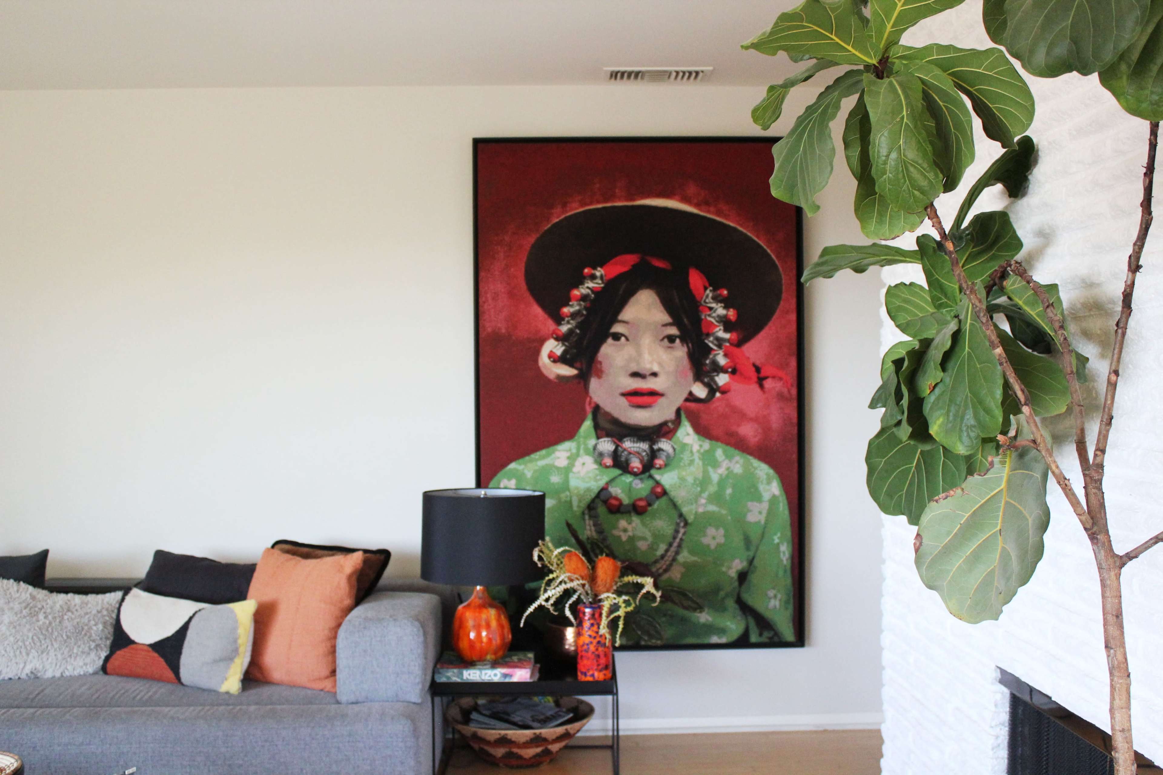 A large portrait of a woman in traditional attire hanging on a wall above a gray sofa with decorative pillows and a plant nearby.