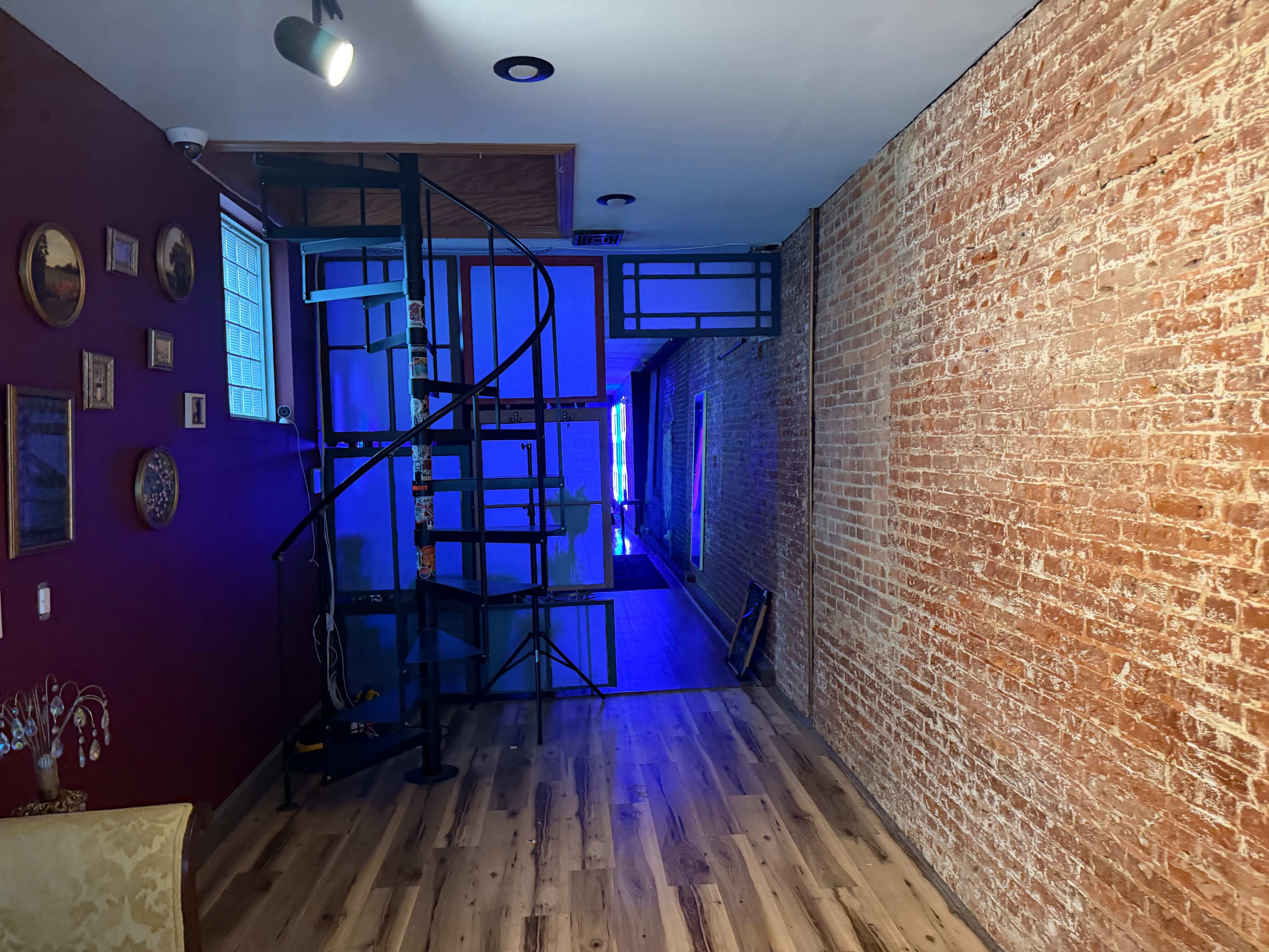 The image shows a room with exposed brick walls, a spiral staircase leading to an upper level, and a brightly lit area at the far end.