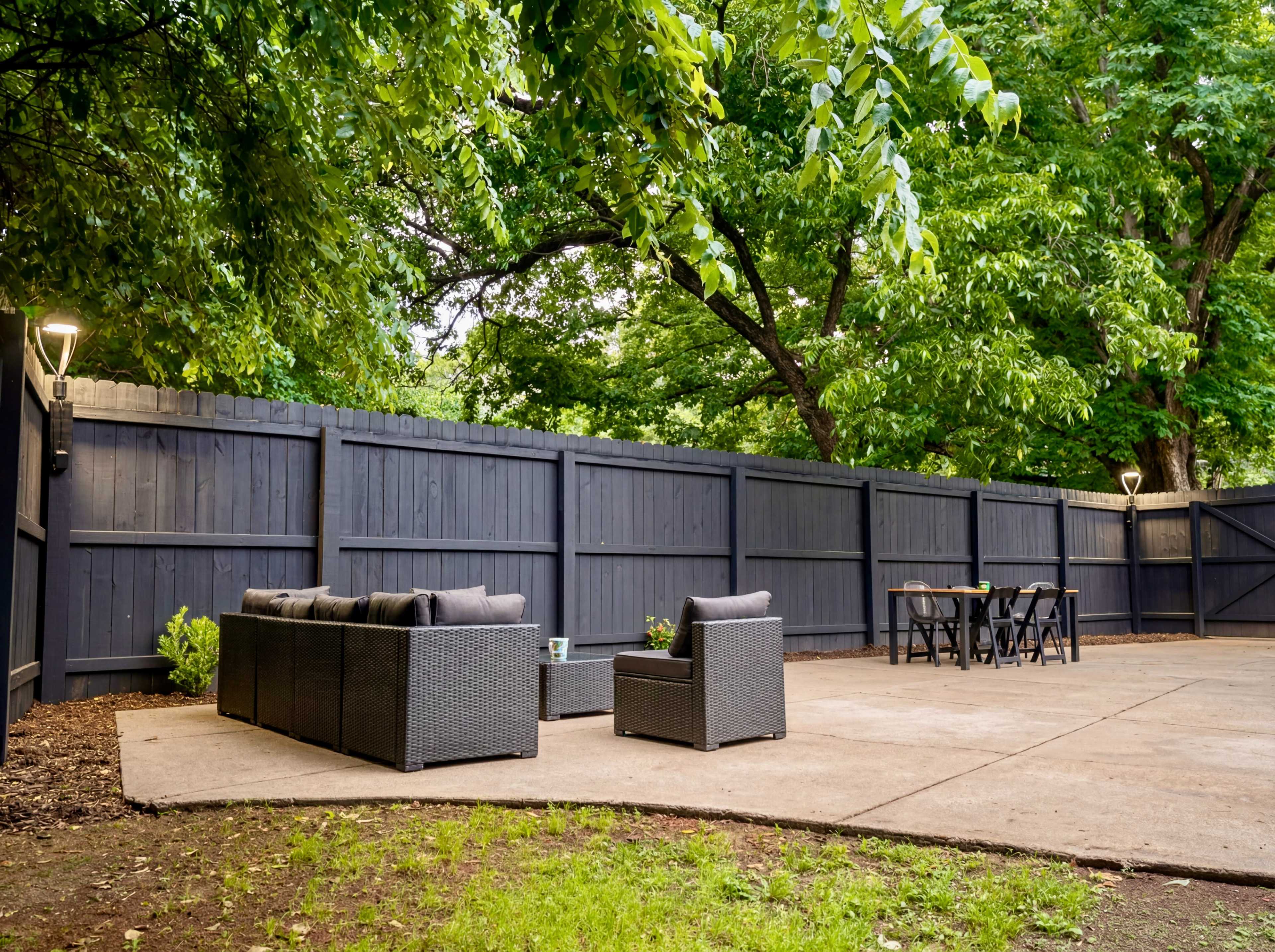 The image shows a patio area with a seating arrangement and a dining table surrounded by a wooden fence and lush green trees.