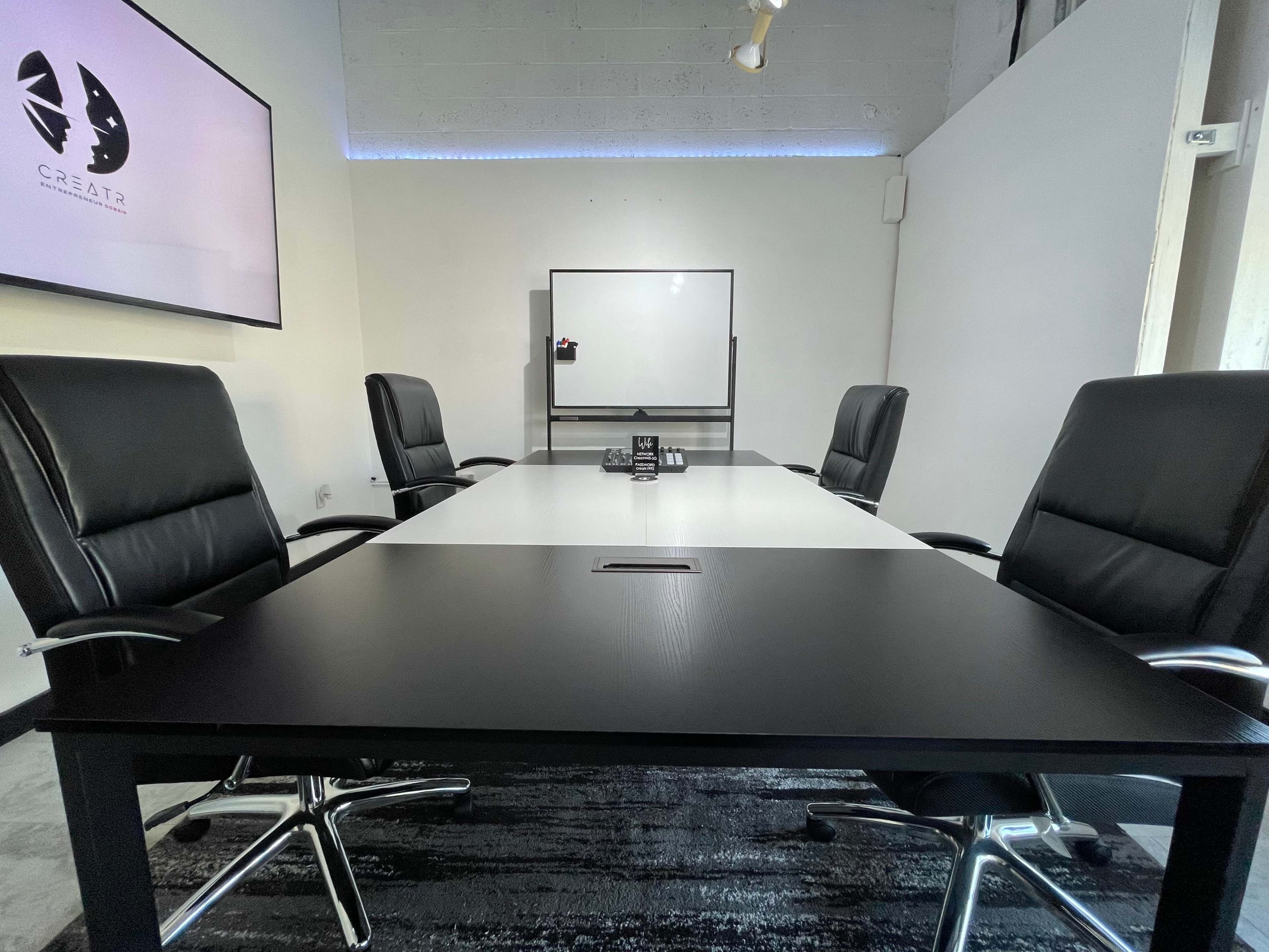 A modern conference room features a large table with black leather chairs and a wall-mounted screen displaying a logo.