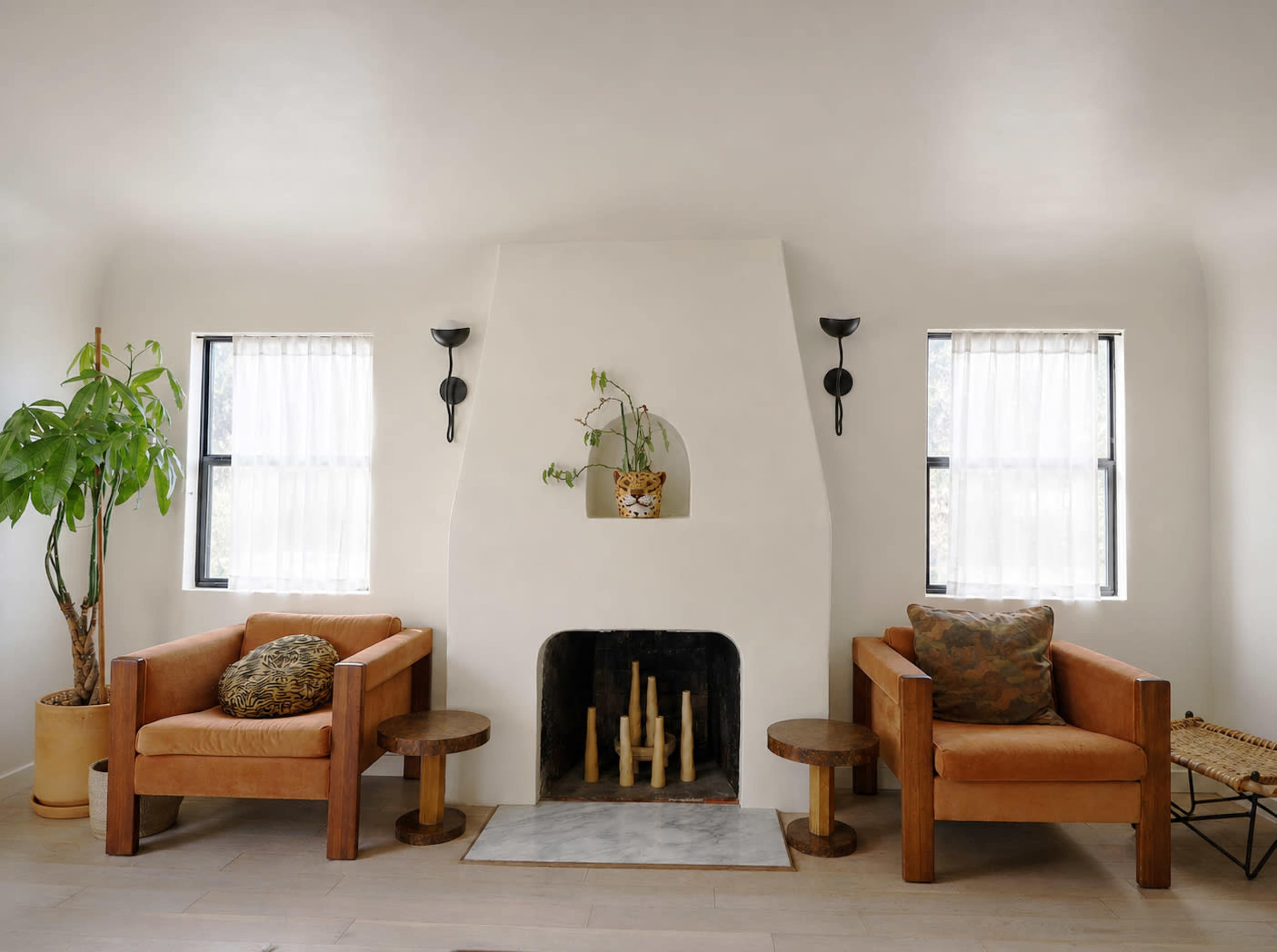 The image features a minimalist living room with two brown armchairs on either side of a white, angular fireplace, flanked by windows with sheer curtains.