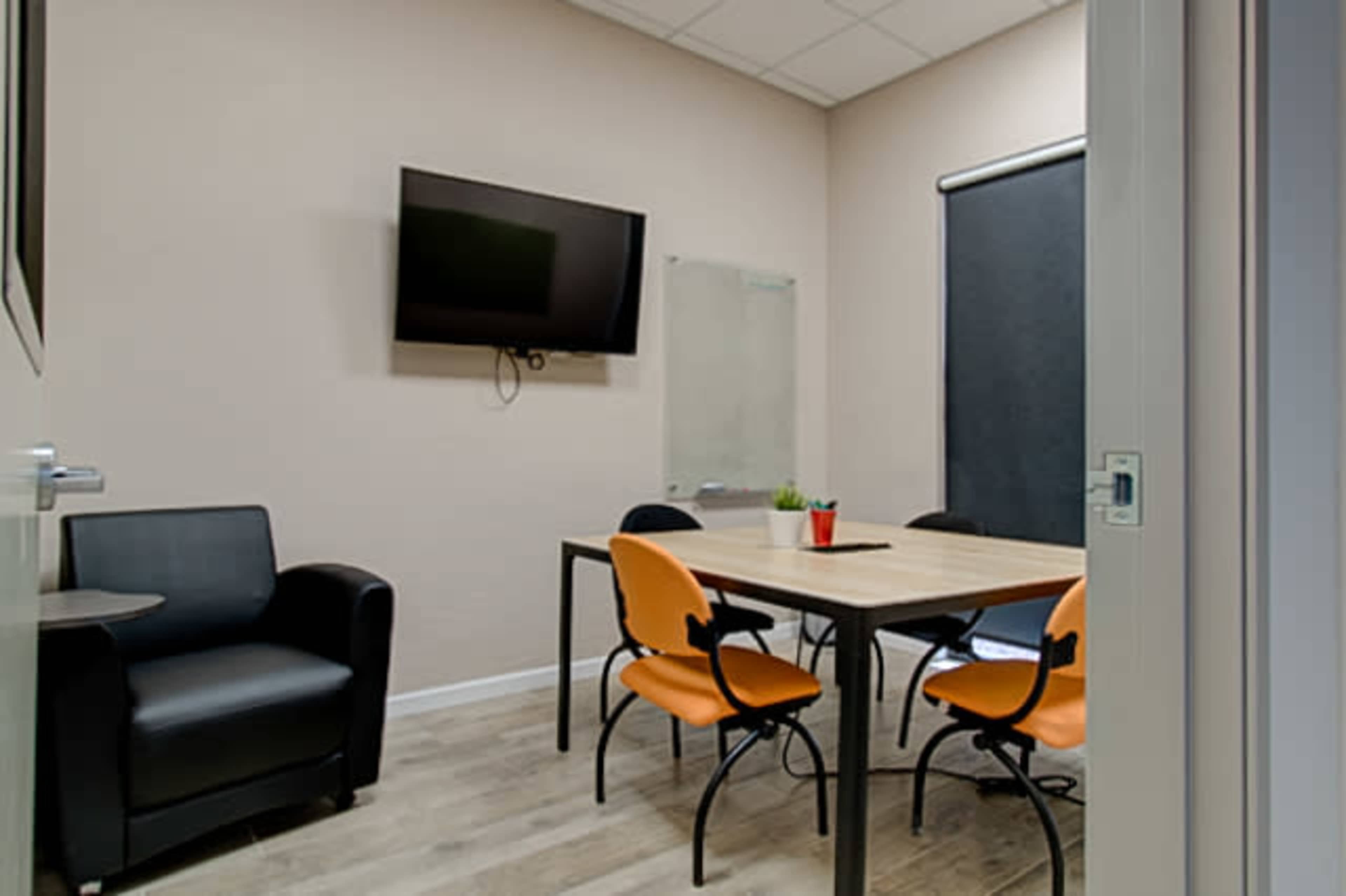 A small conference room features a table surrounded by black chairs and orange chairs, with a television mounted on the wall and a window covered by a black shade.