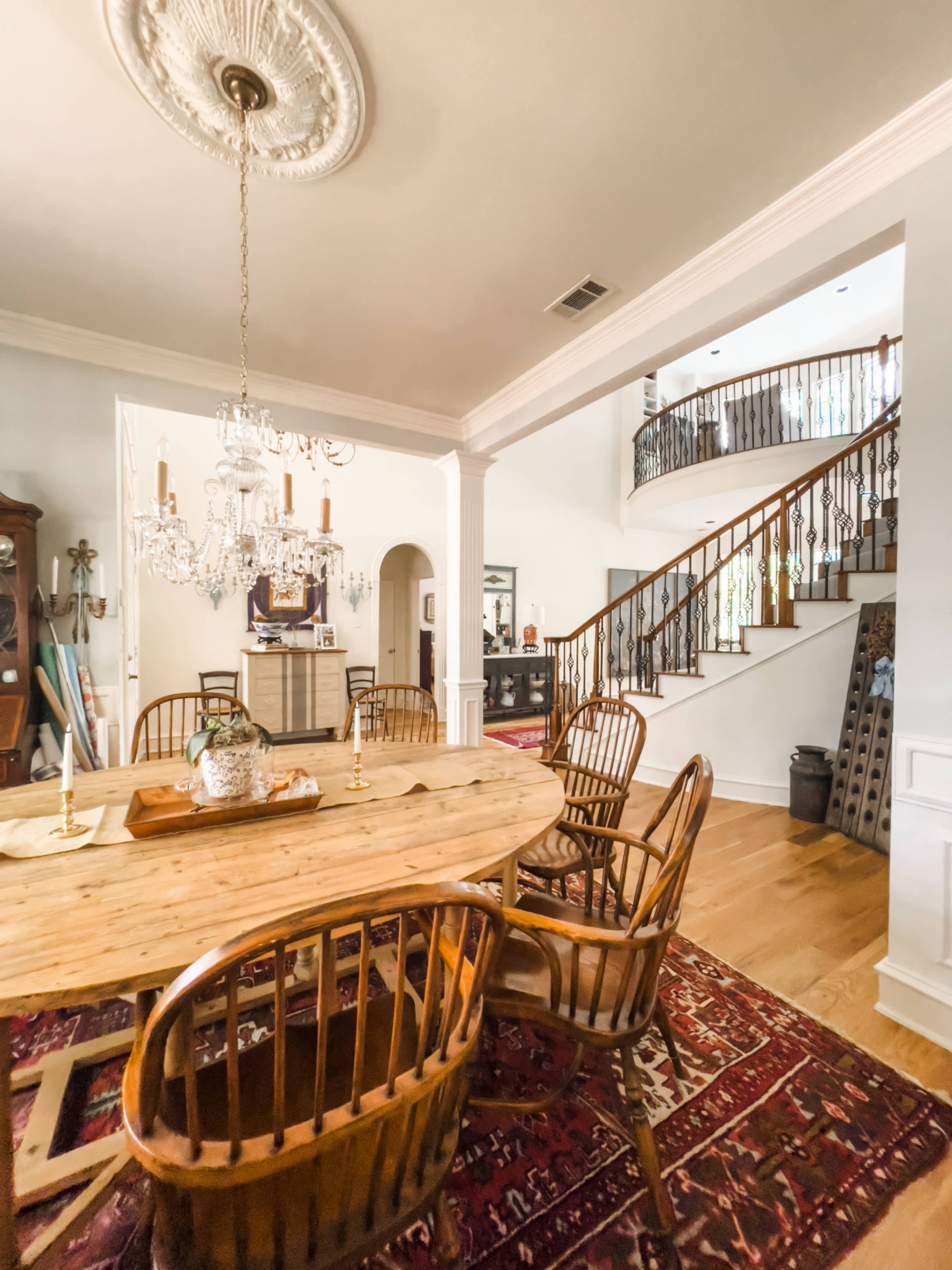 The image shows a dining area with a large wooden table surrounded by chairs, a chandelier above, and an open staircase leading to a second floor in a well-lit room.