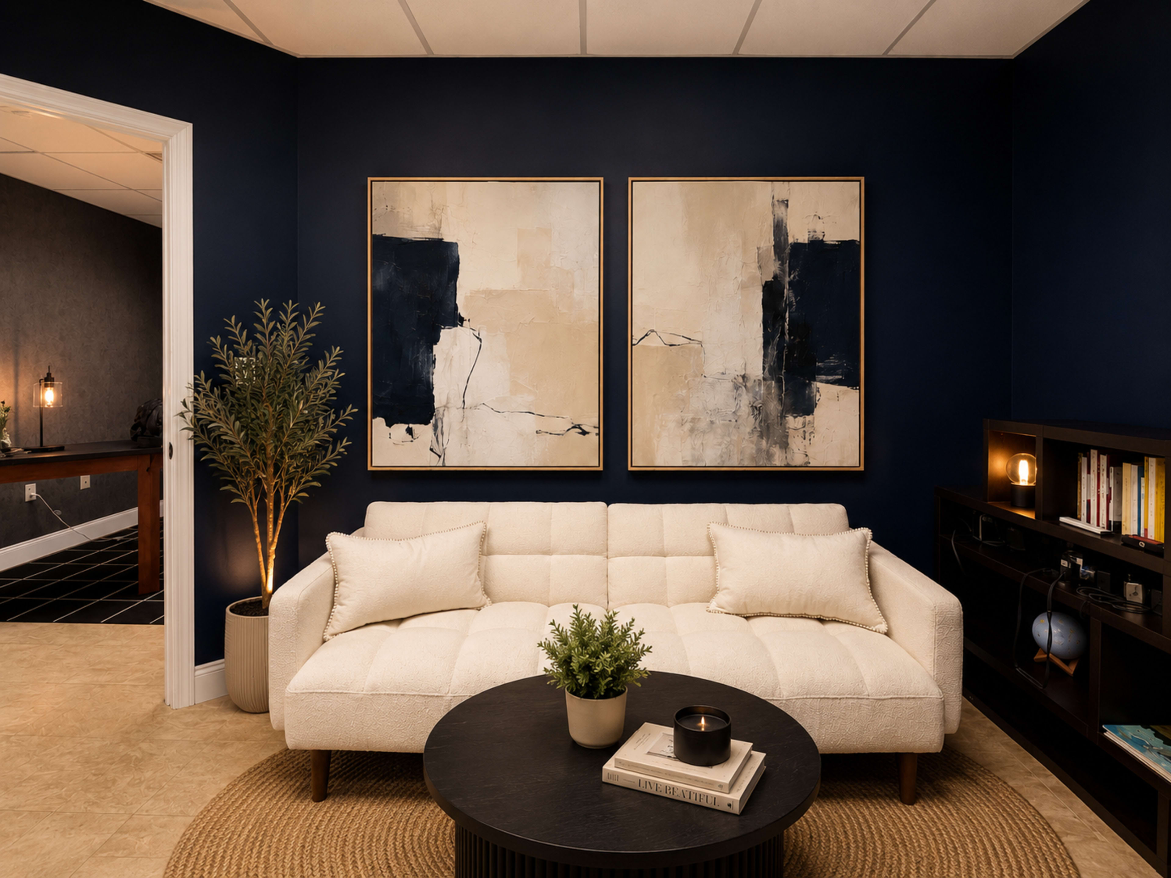 A modern living room features a light-colored sofa, two abstract paintings on a dark blue wall, a round black coffee table, and a decorative plant.