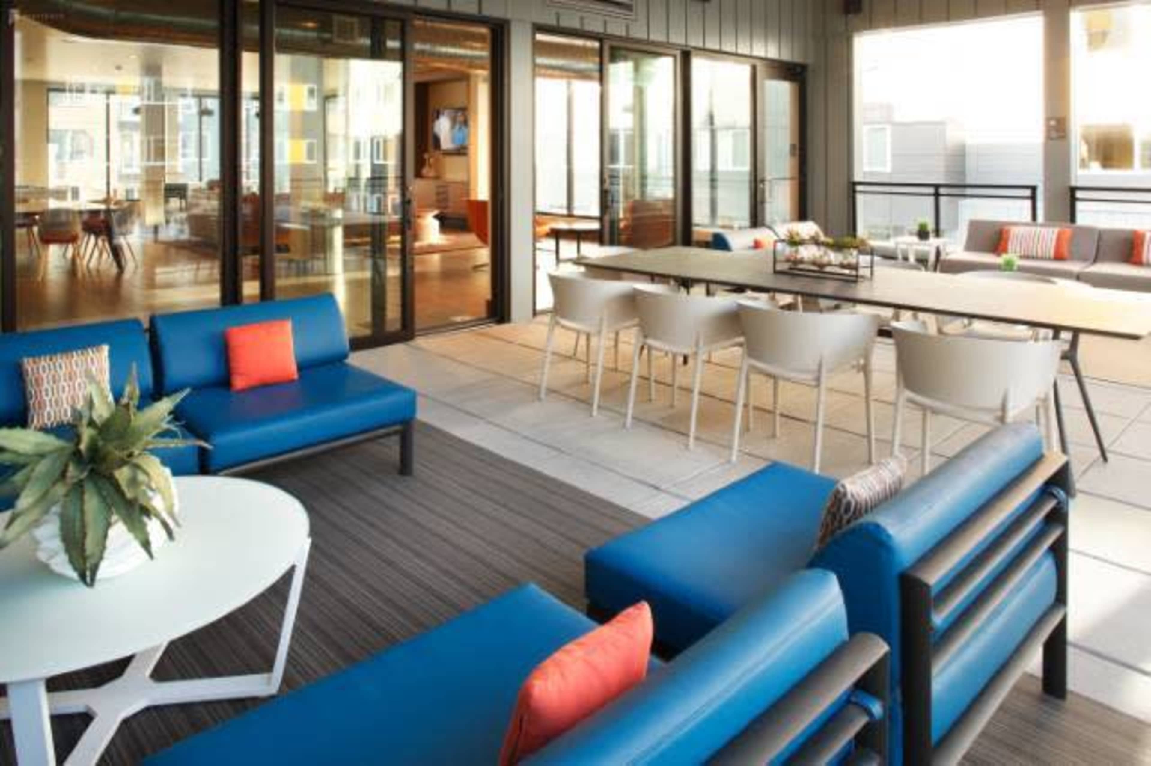 A modern outdoor lounge area with blue seating, a coffee table, and a dining table surrounded by chairs, all situated near large glass doors leading into an indoor space.