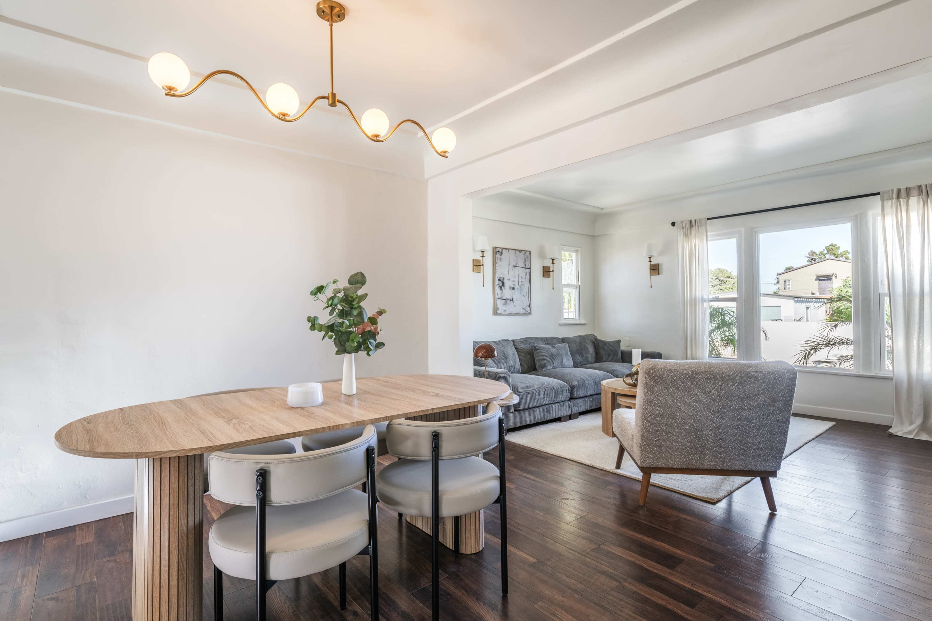 The image shows a modern dining area with a round wooden table and four chairs, adjacent to a cozy living space featuring a gray sectional sofa and large windows.