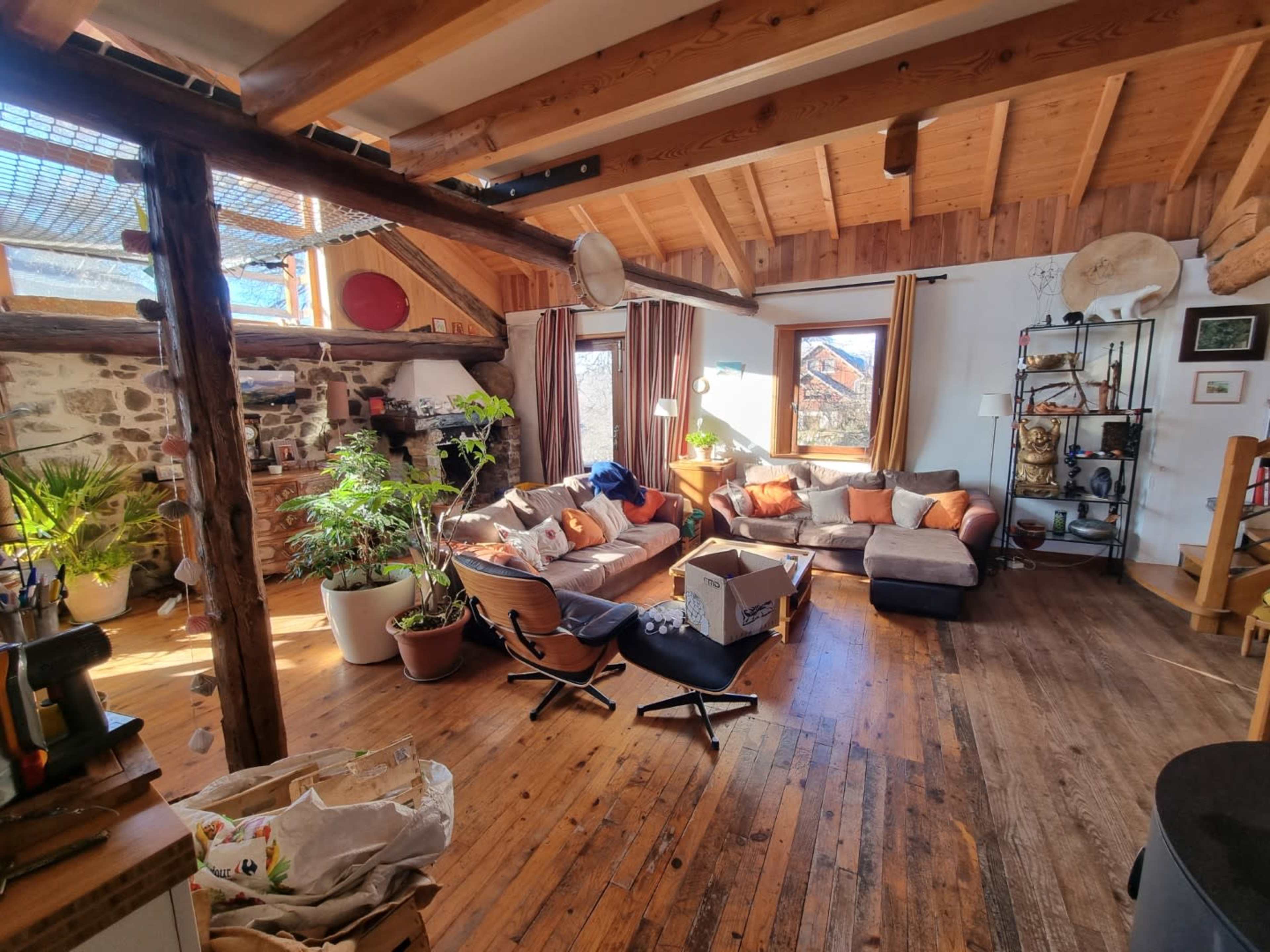 A spacious living room features a large sectional sofa with colorful cushions, wooden beams, and plants, along with a mix of wooden and modern furniture.