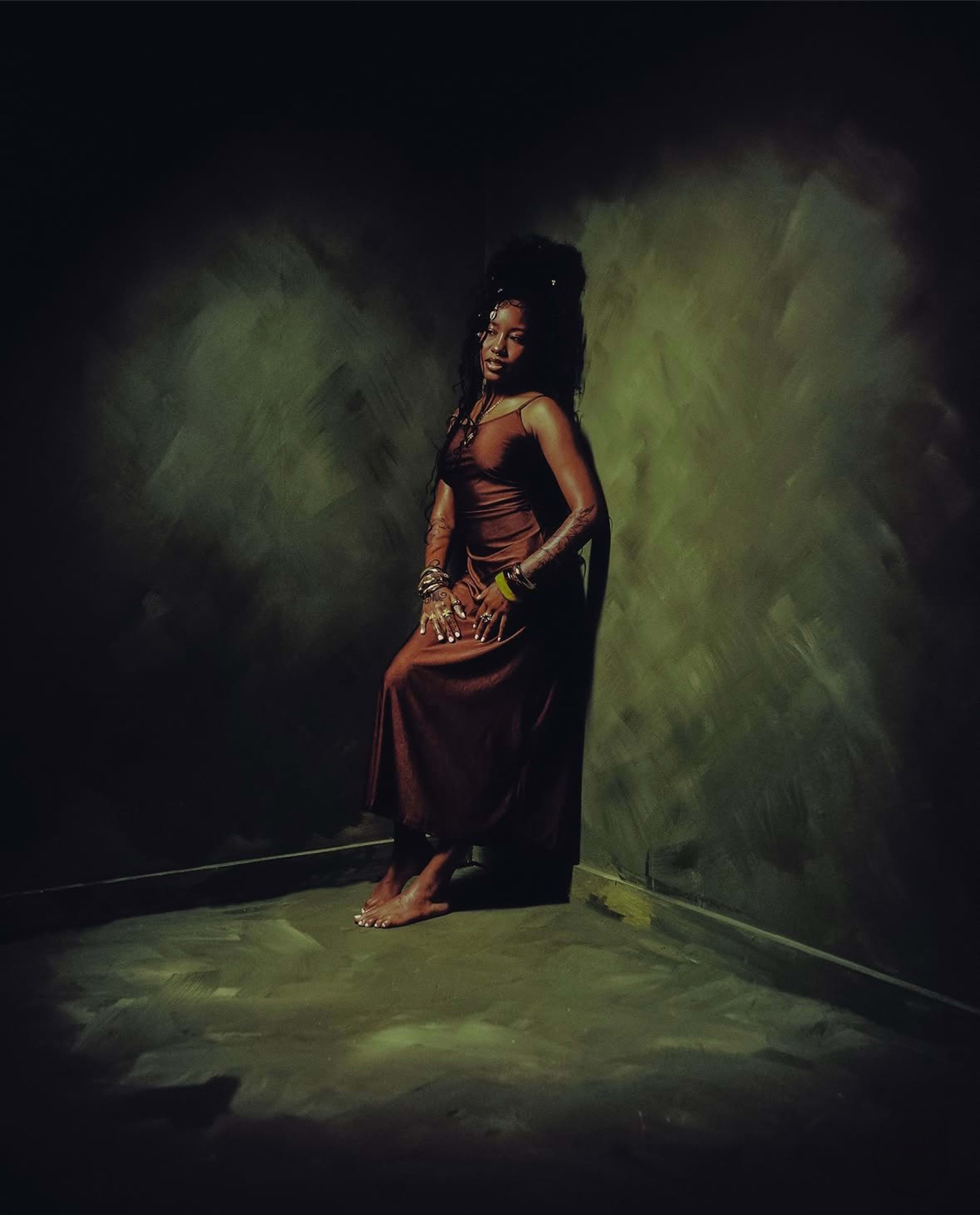 A woman in a brown dress sits against a textured green wall in a dimly lit corner.