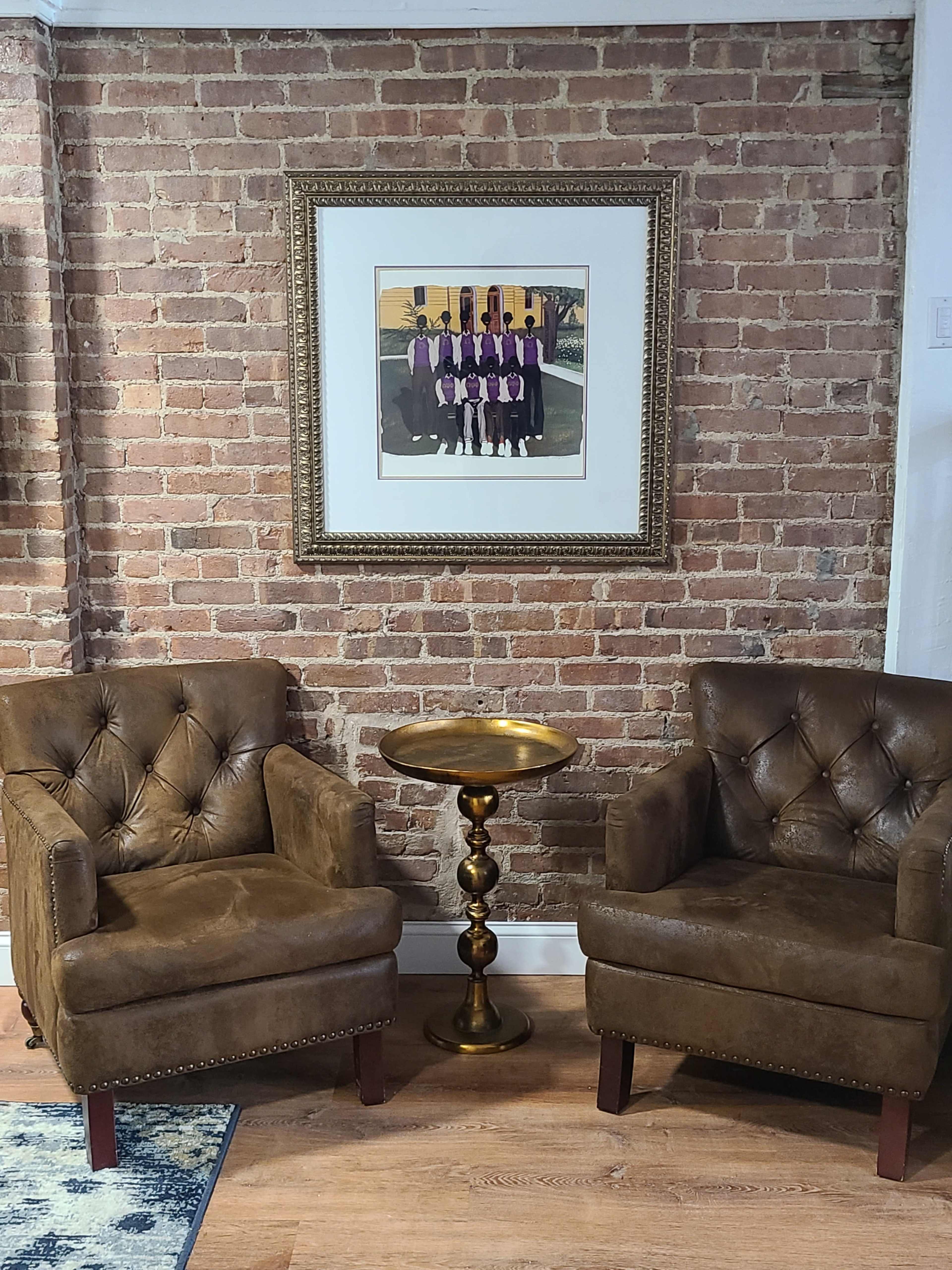 Two brown, tufted armchairs are positioned on either side of a small, round brass side table in front of a framed artwork hanging on a brick wall.