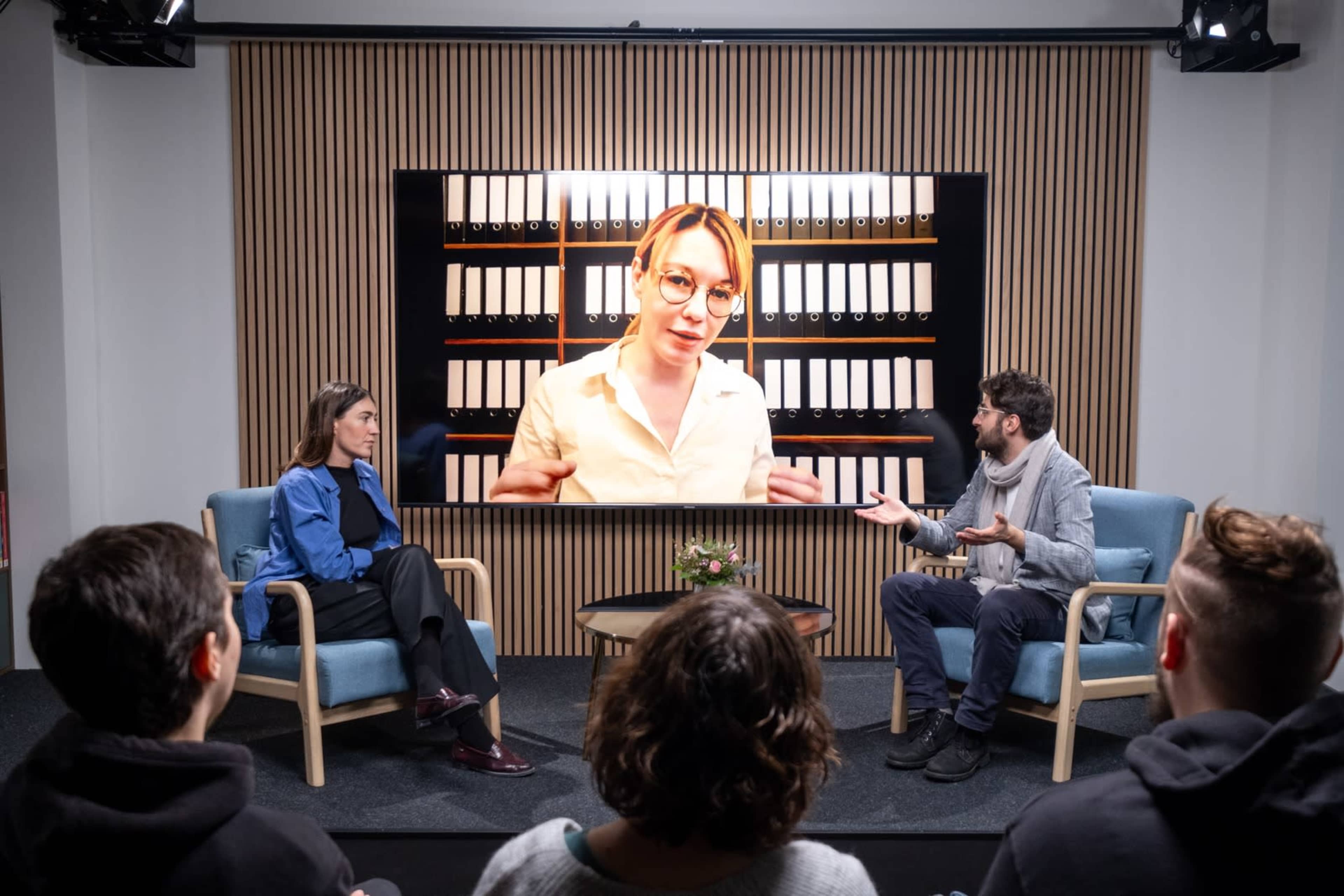 A virtual discussion takes place with two people seated in chairs facing a large screen displaying a person talking.