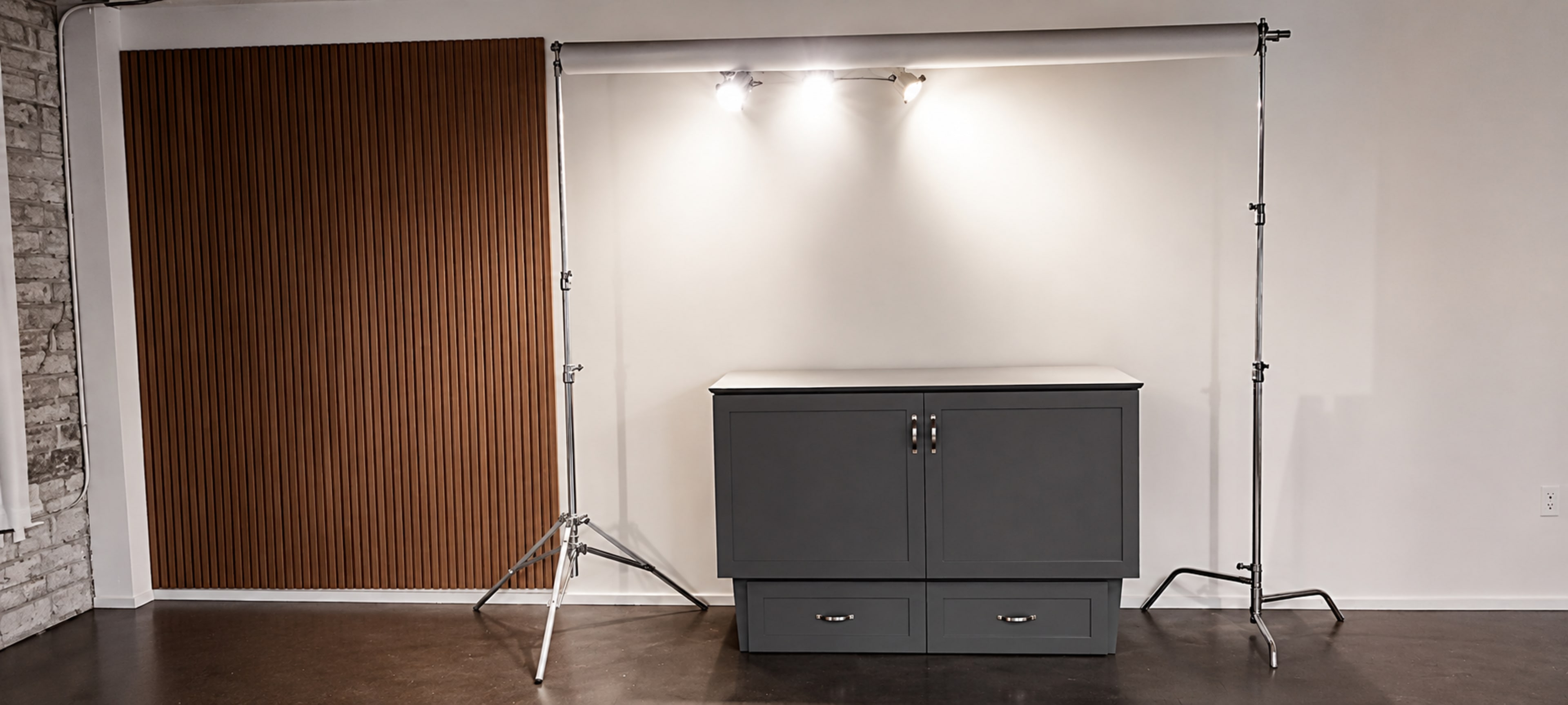 The image shows a minimalist studio setup with a gray cabinet and a backdrop support frame fitted with two lights against a plain wall.