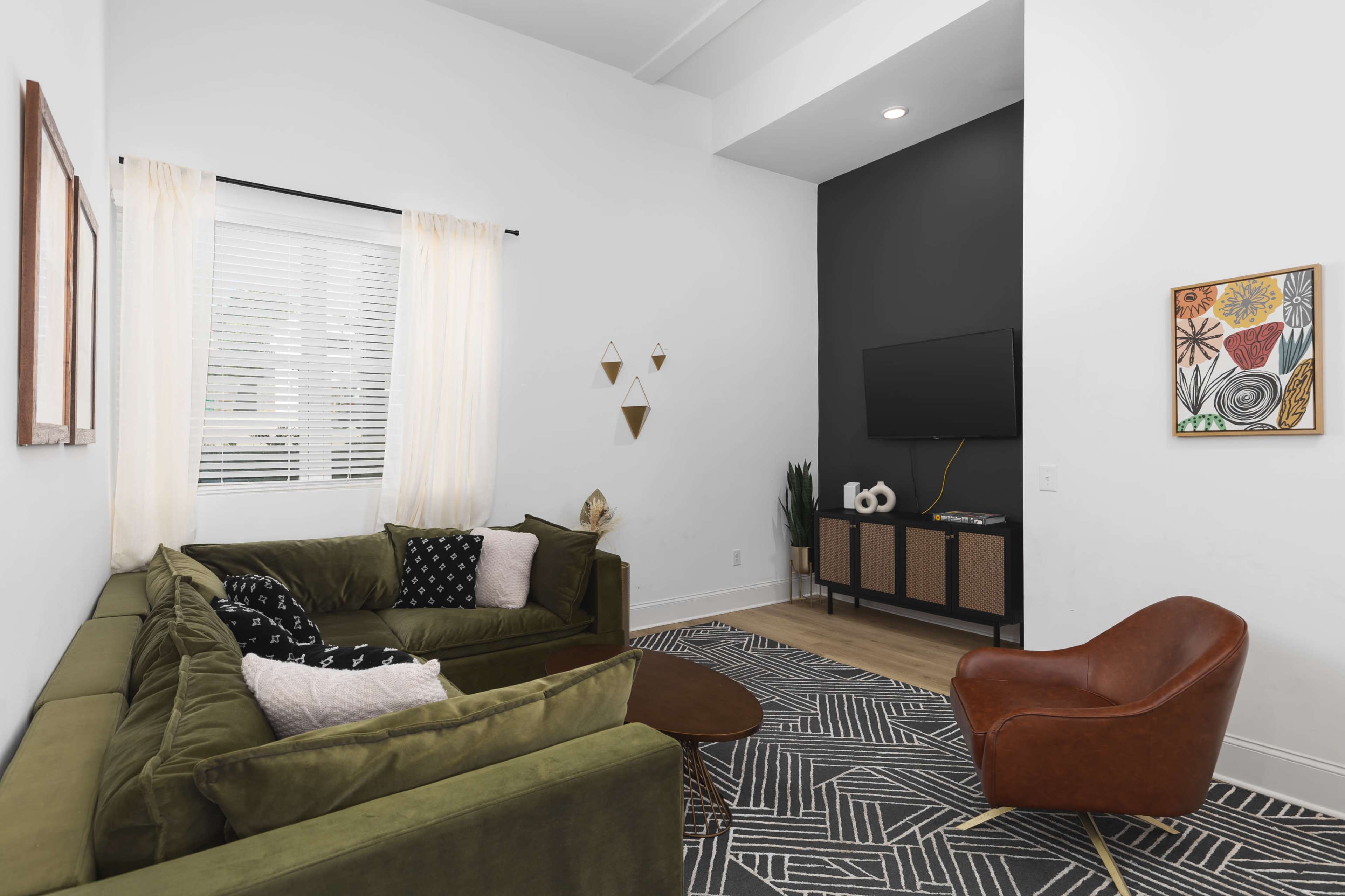 A modern living room features a green sectional sofa, a round coffee table, and a brown accent chair, with a TV mounted on a dark accent wall and decorative art pieces around the space.