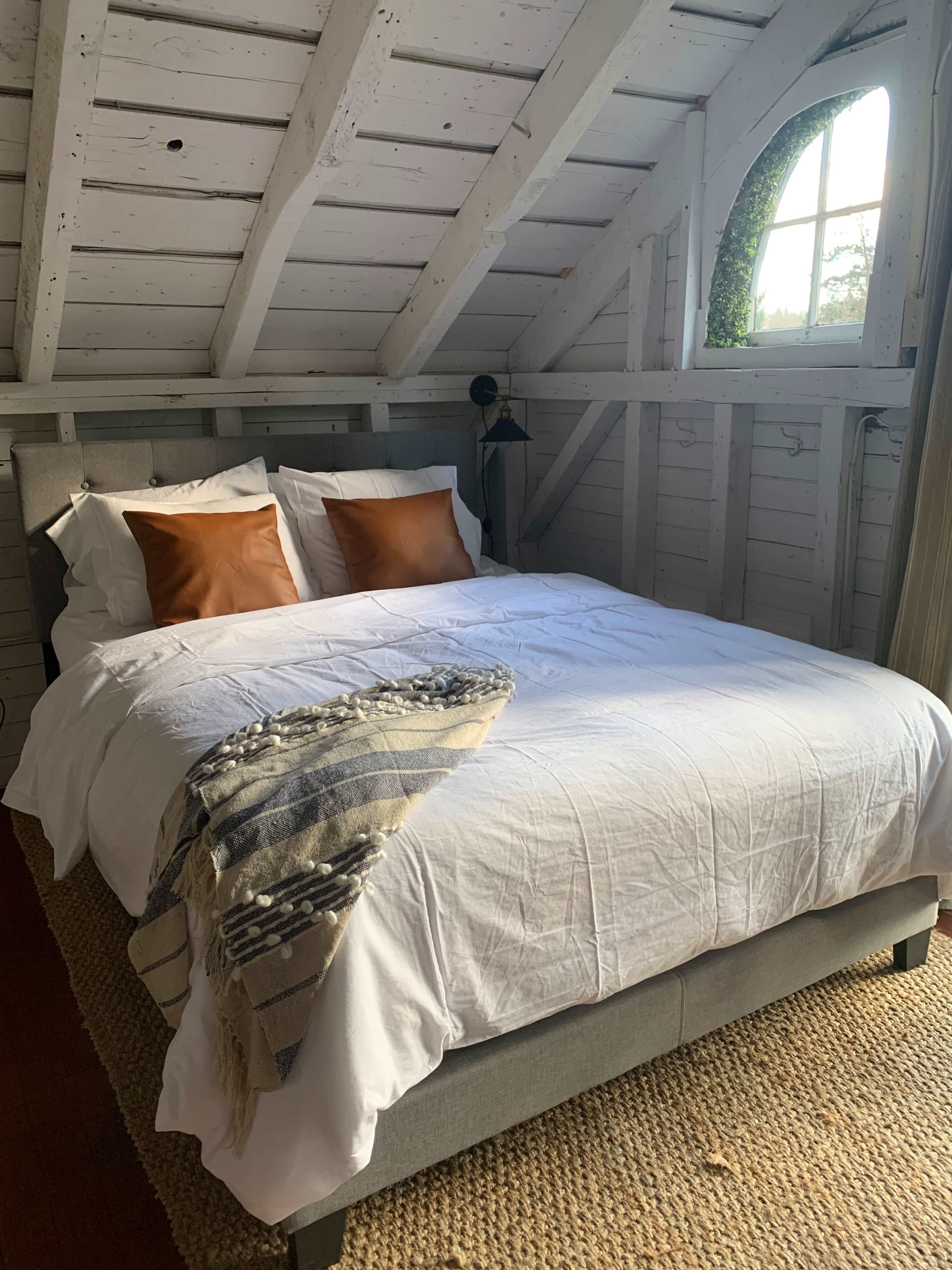The image shows a neatly made bed with white linens and two orange pillows in a cozy attic room featuring wooden beams and a window.